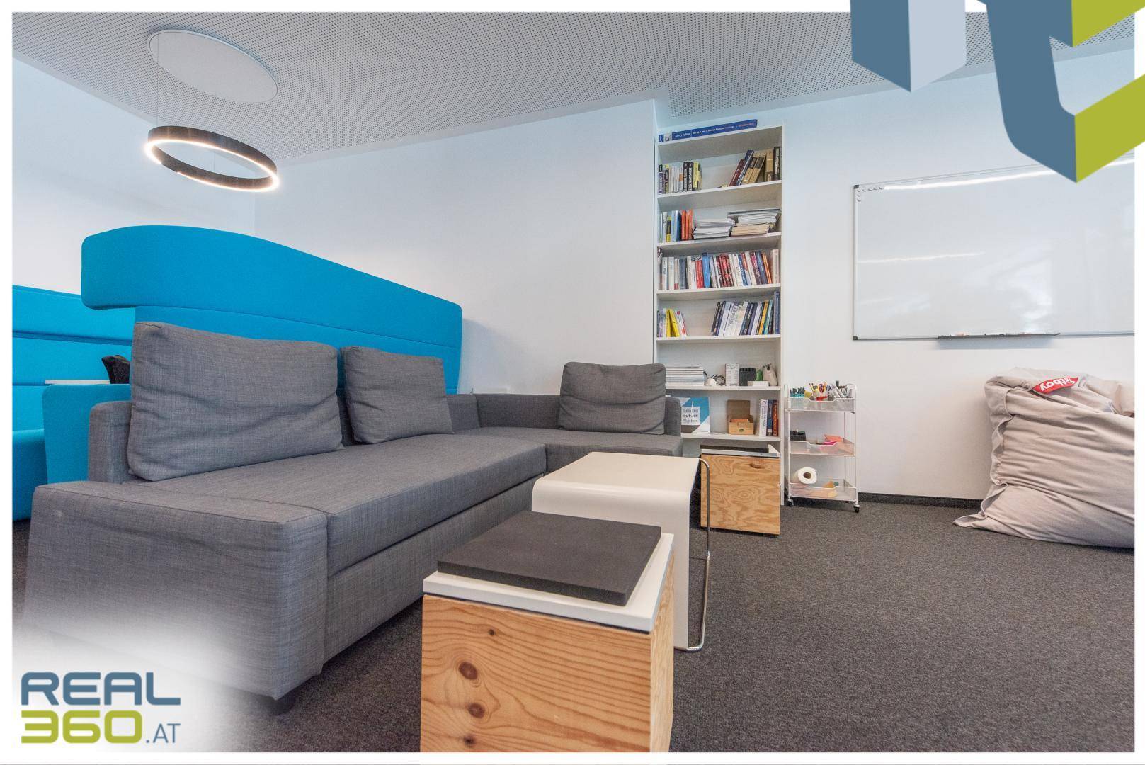Entspannter Loungebereich mit grauem Sofa, Bücherregal und Whiteboard für kreative Pausen.