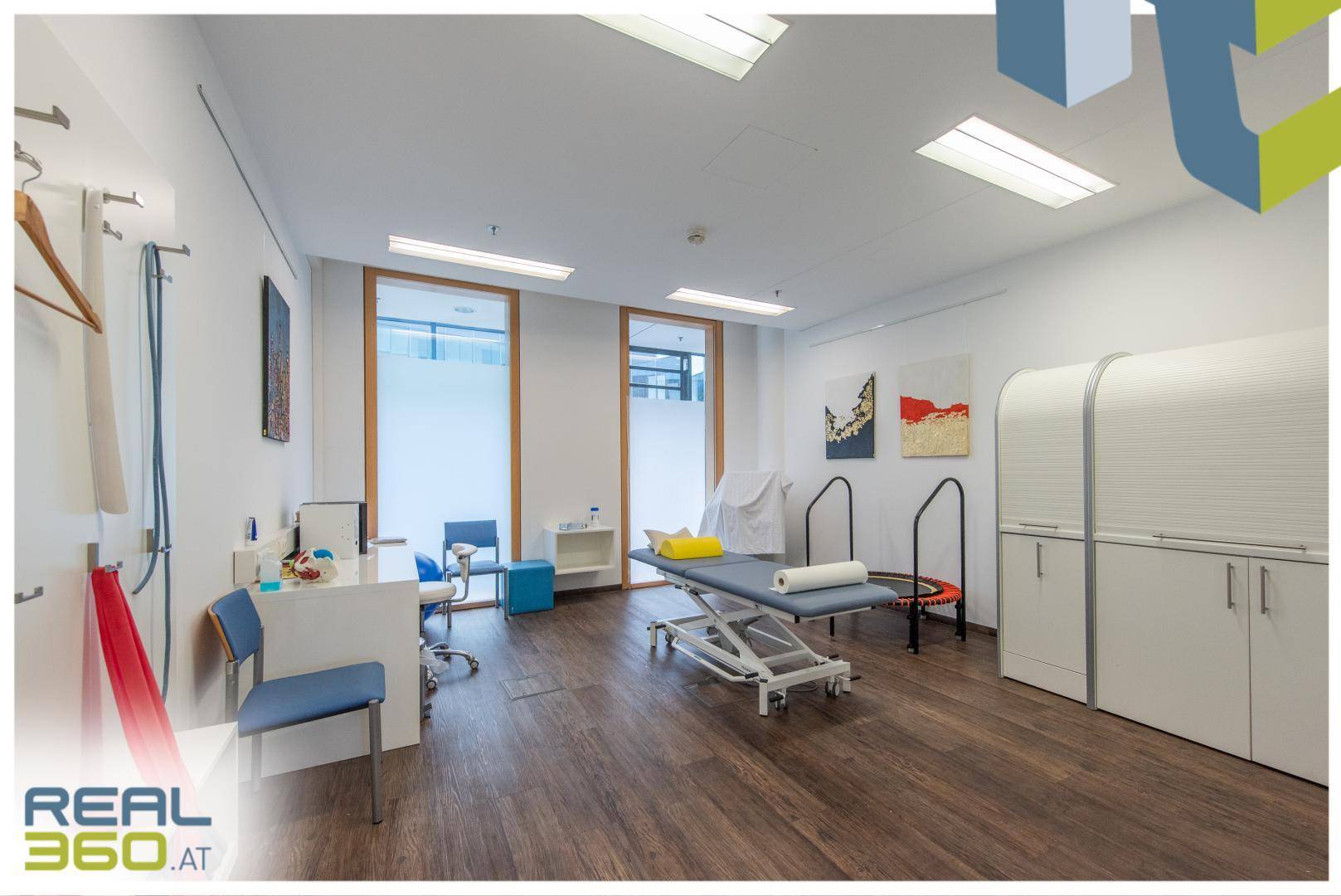 Heller Therapieraum mit Holzboden, Behandlungsliege und verschiedenen Trainingsgeräten, gut beleuchtet.