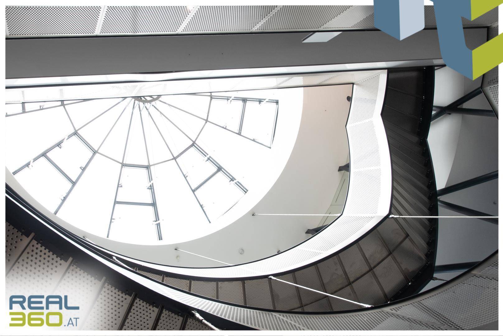 Blick nach oben auf eine beeindruckende Glaskuppel und die geschwungene Metalltreppe in einem modernen Gebäude.