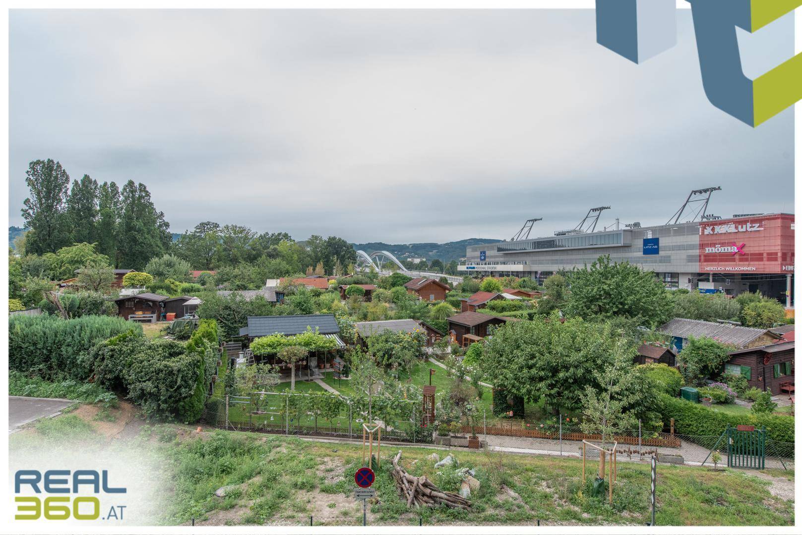 Weitläufiger Blick auf eine Kleingartenanlage mit Häusern und einem Einkaufszentrum im Hintergrund.