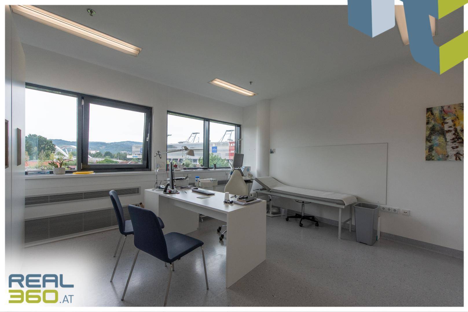 Helles Untersuchungszimmer mit Schreibtisch, Stühlen, Liege und großen Fenstern mit Ausblick.