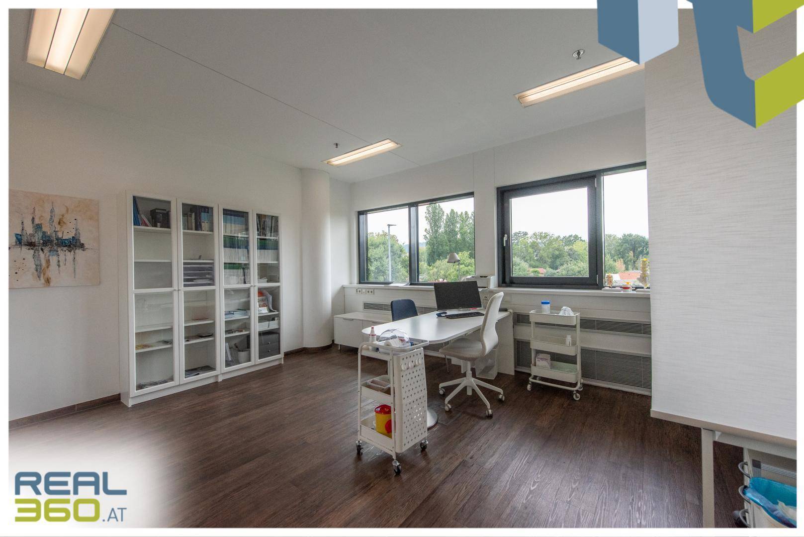 Geräumiges Büro oder Behandlungszimmer mit Holzboden, Schreibtisch, Regalen und Fenstern mit Blick ins Grüne.