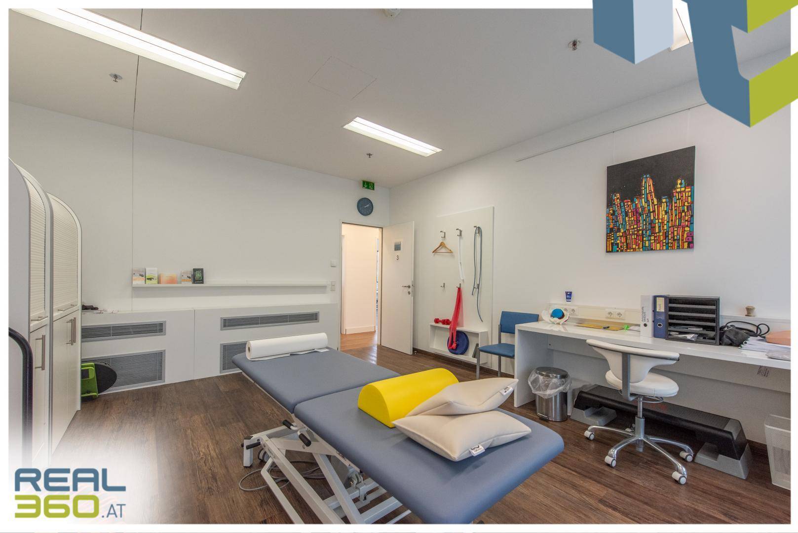 Therapieraum mit Behandlungsliege, Schreibtisch und Kunst an der Wand, funktional und hell gestaltet.