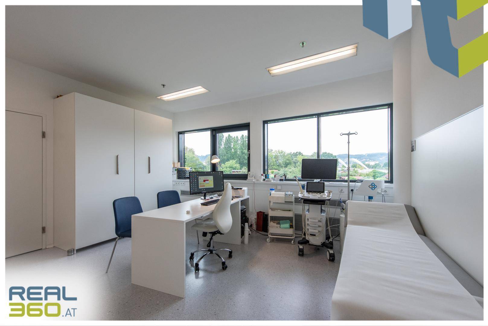 Helles Untersuchungszimmer mit Schreibtisch, Liege und medizinischen Geräten, große Fenster bieten Ausblick.
