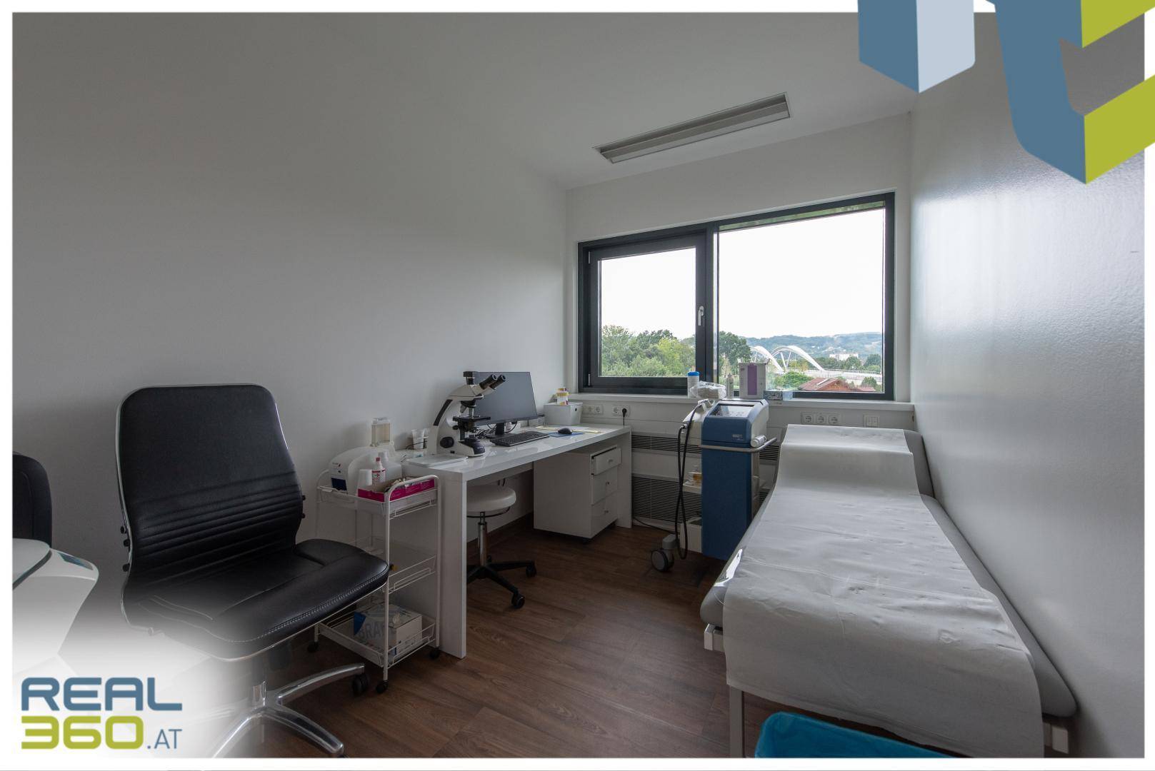 Helles Untersuchungszimmer mit Holzboden, einem Schreibtisch, Liege und Fenstern mit Blick ins Grüne.