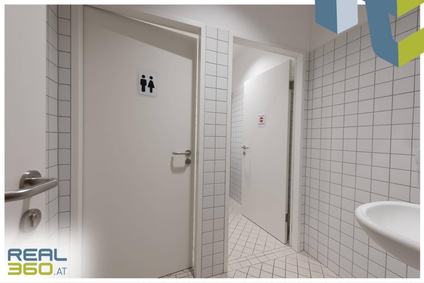 Saubere Toilettenräume mit weißen Fliesen, Waschbecken und klaren Beschilderungen an den Türen.
