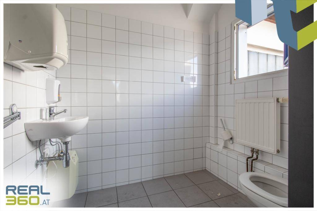 Einfache Toilette mit weißen Fliesen, Waschbecken, WC und Fenster für natürliche Belüftung.