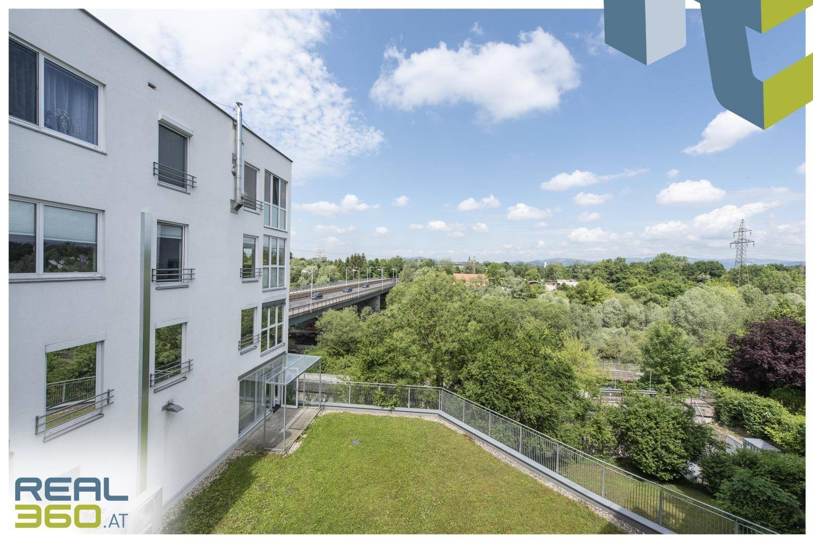 Weitläufige Dachterrasse mit Rasenfläche und Panoramablick auf die grüne Landschaft und eine Brücke.