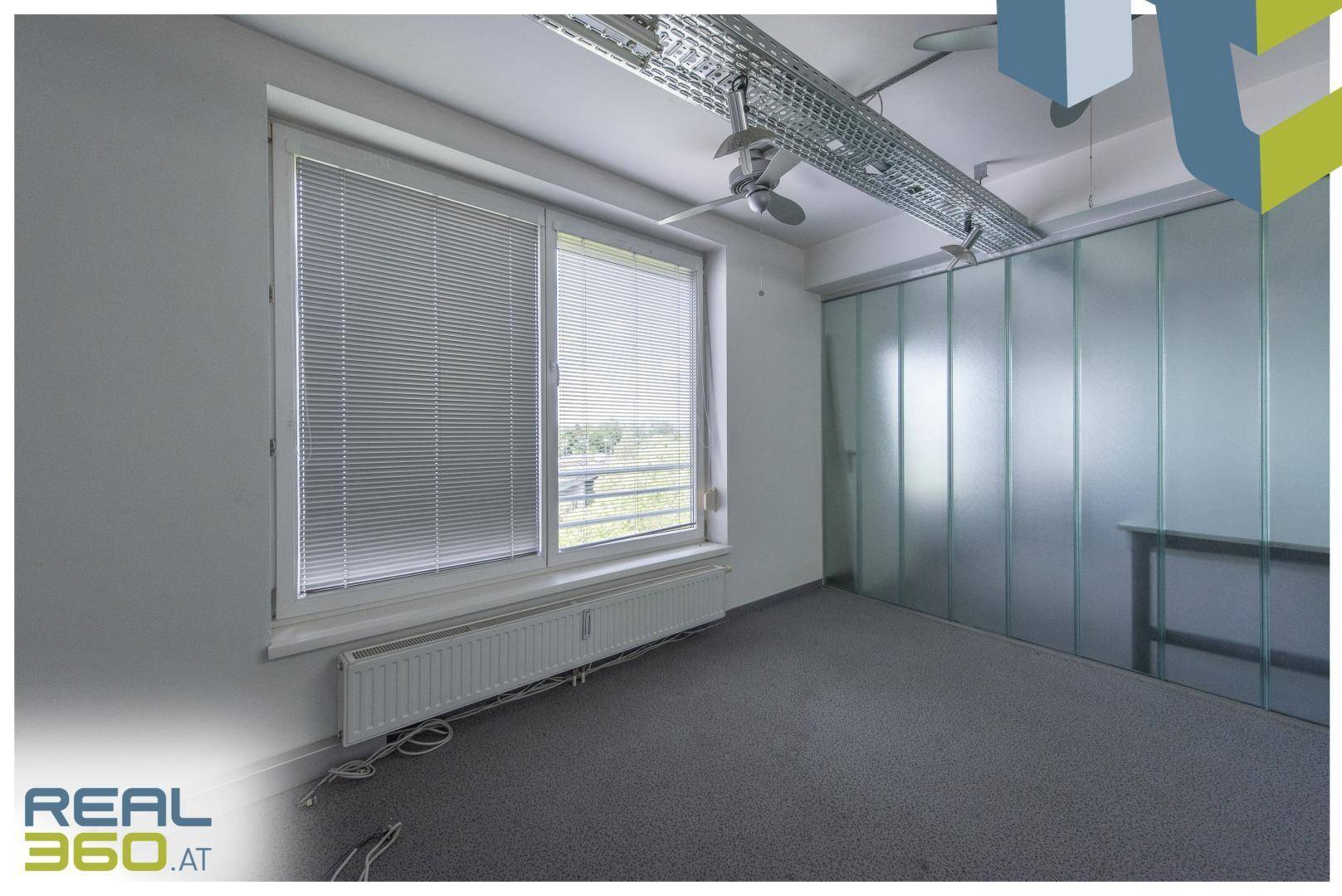 Heller Büroraum mit Teppichboden, großen Fenstern mit Jalousien und einer modernen Glaswand.