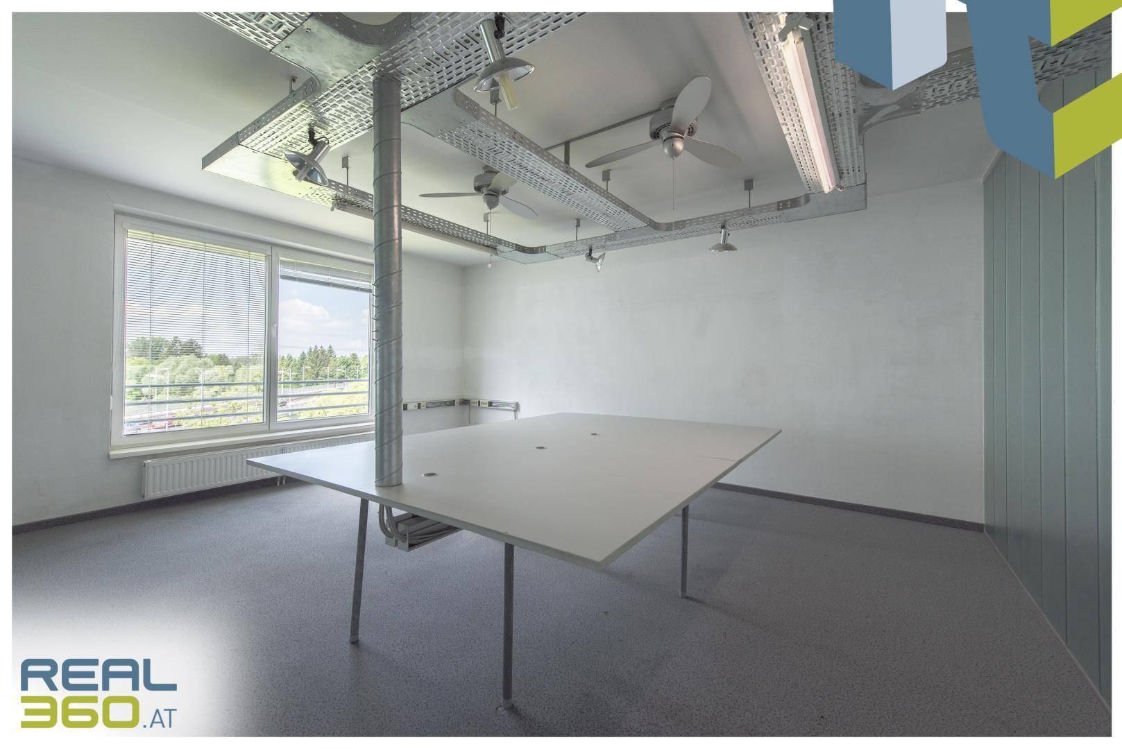 Offener Bürobereich mit einem großen Arbeitstisch, Blick ins Grüne und sichtbaren Deckeninstallationen.