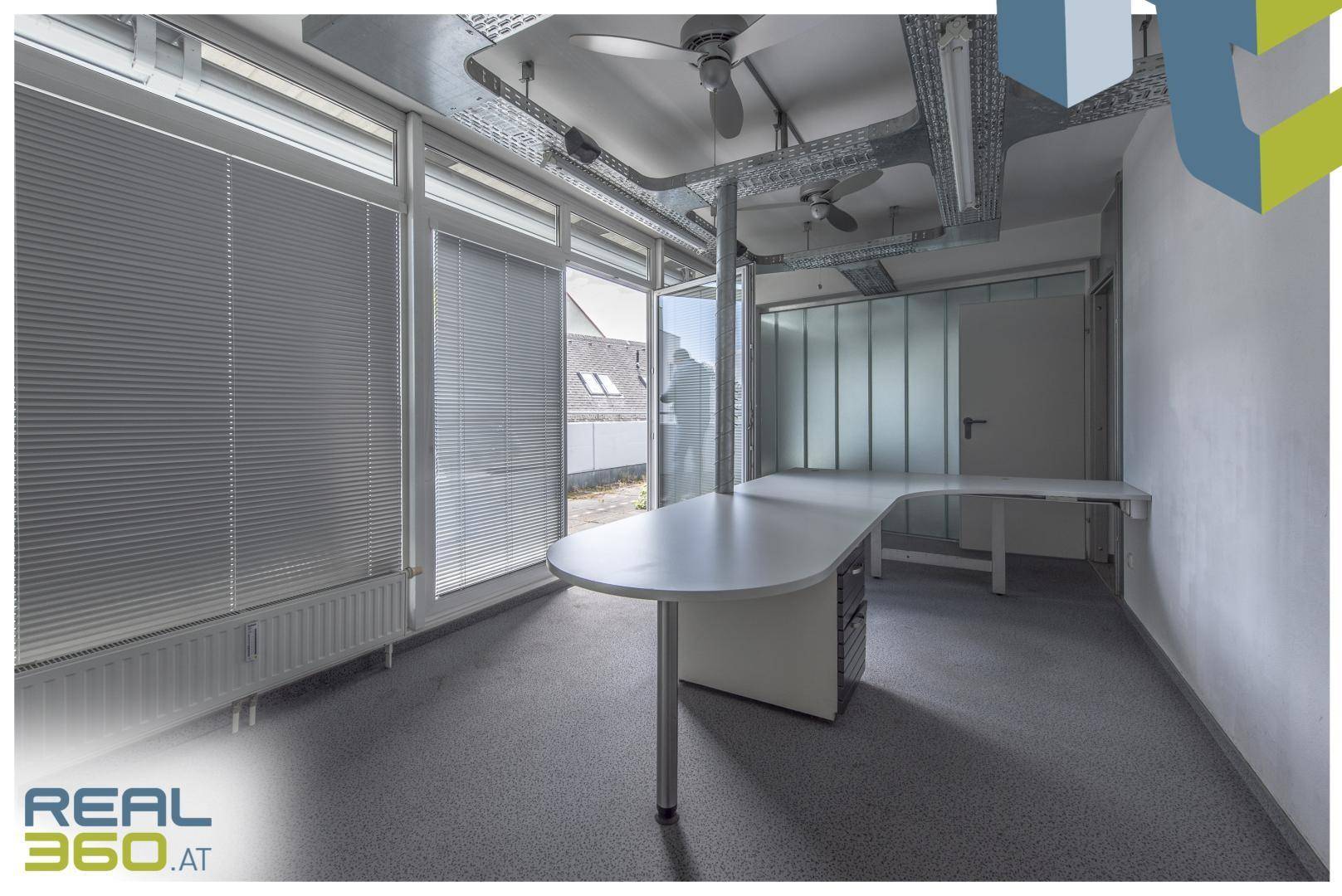 Heller Büroraum mit L-förmigem Schreibtisch, Jalousien an den Fenstern und industriellen Deckeninstallationen.