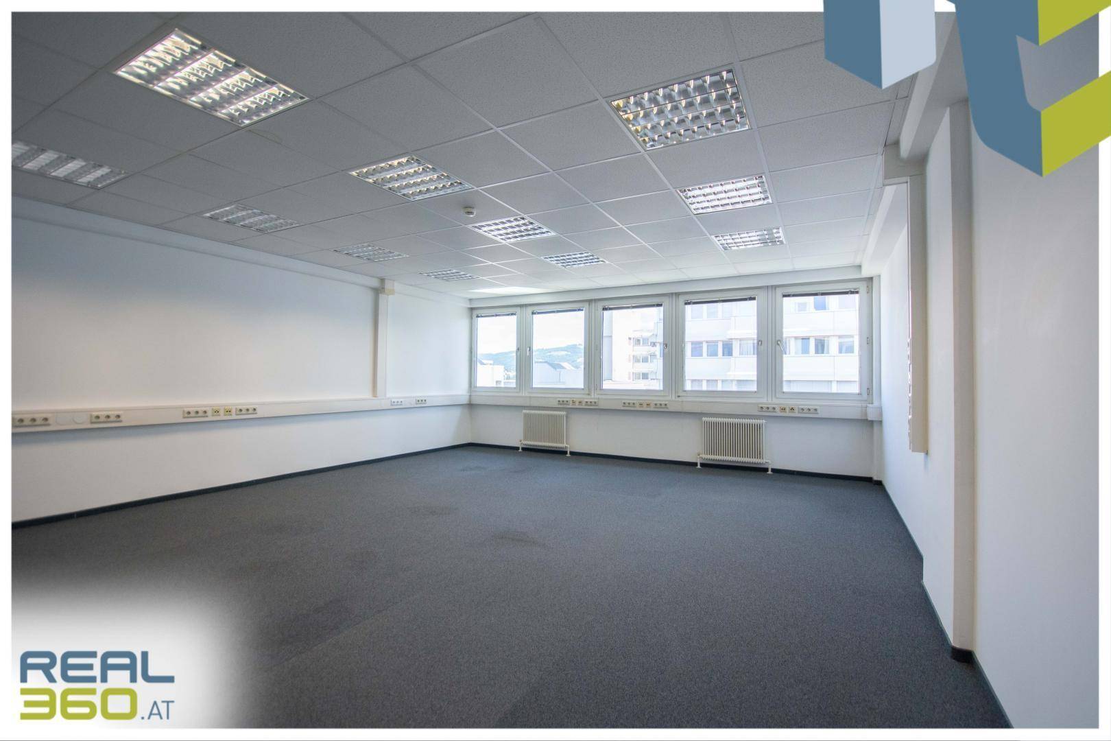 Geräumiges Büro mit großen Fenstern, Teppichboden und abgehängter Decke, gut beleuchtet.