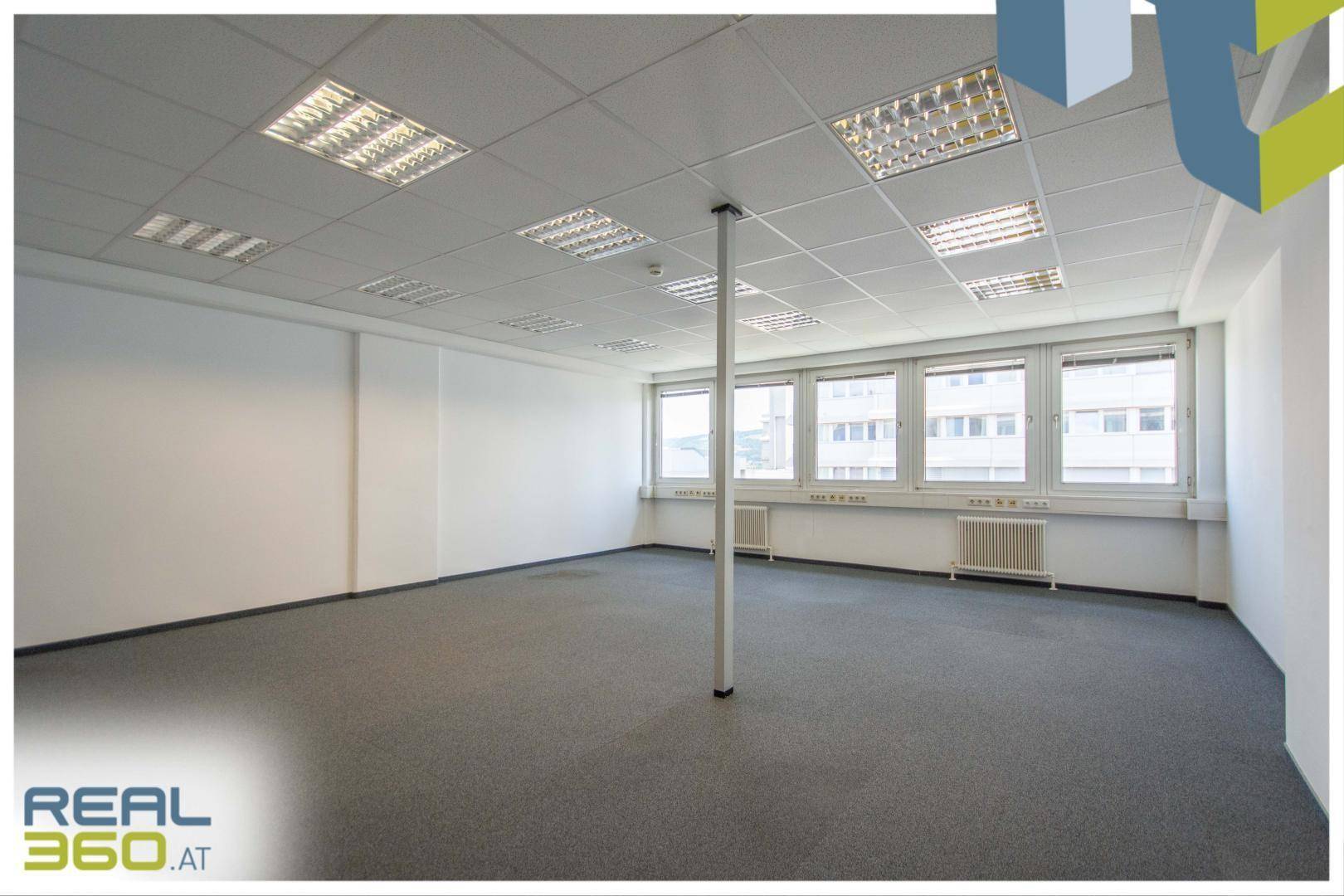 Geräumiger, leerer Büroraum mit Teppichboden, großen Fenstern und abgehängter Decke.
