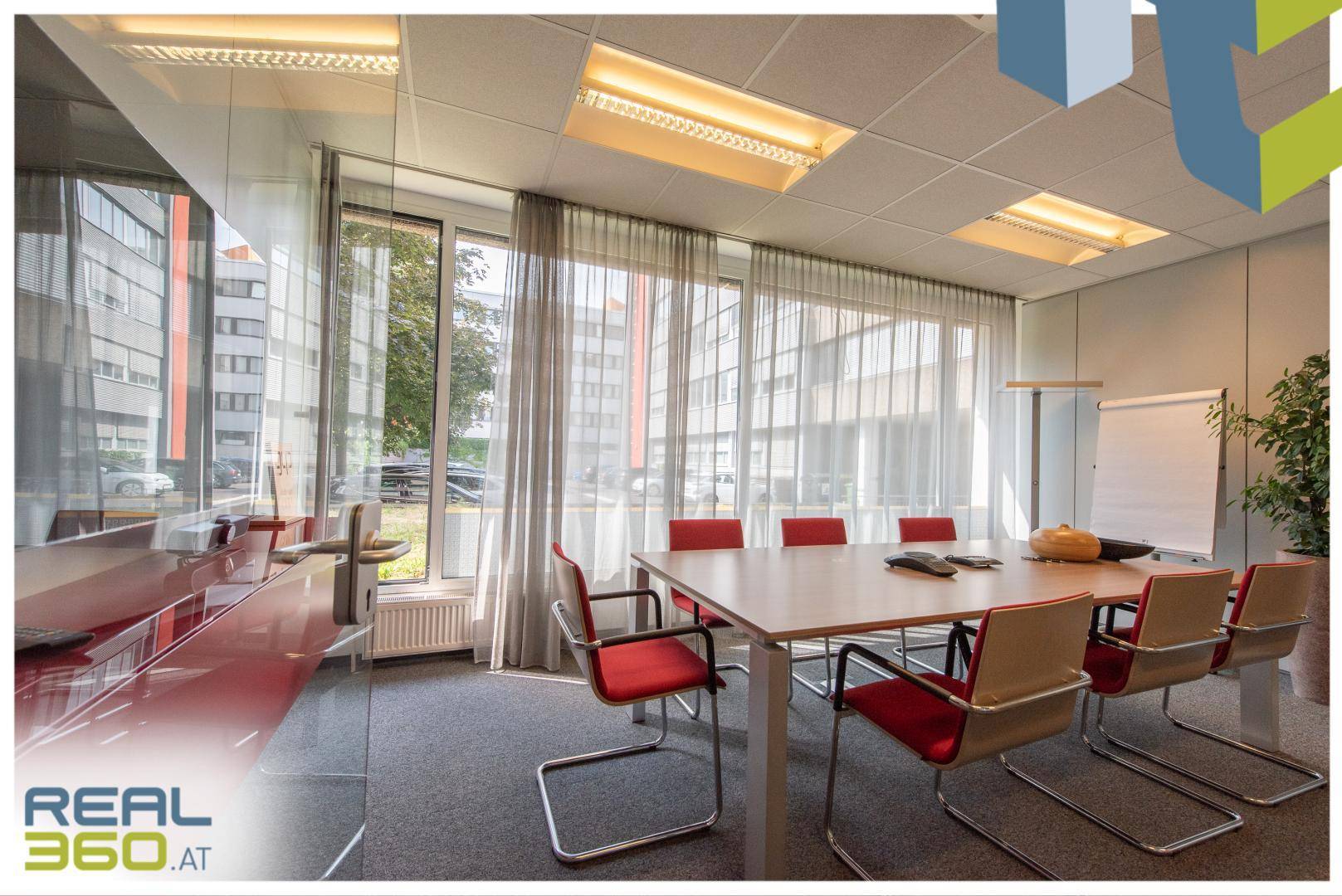 Heller Konferenzraum mit großem Tisch, roten Stühlen und Whiteboard, umgeben von großen Fenstern.