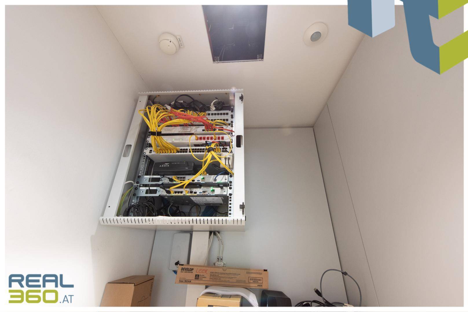 Kleiner Serverraum mit einem Rack voller elektronischer Geräte und Kabel, für die Netzwerkverwaltung.