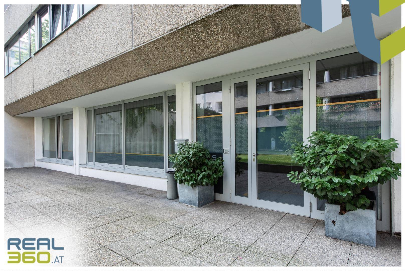 Eingangsbereich eines Bürogebäudes mit Glastüren, gepflasterter Terrasse und dekorativen Pflanzenkübeln.