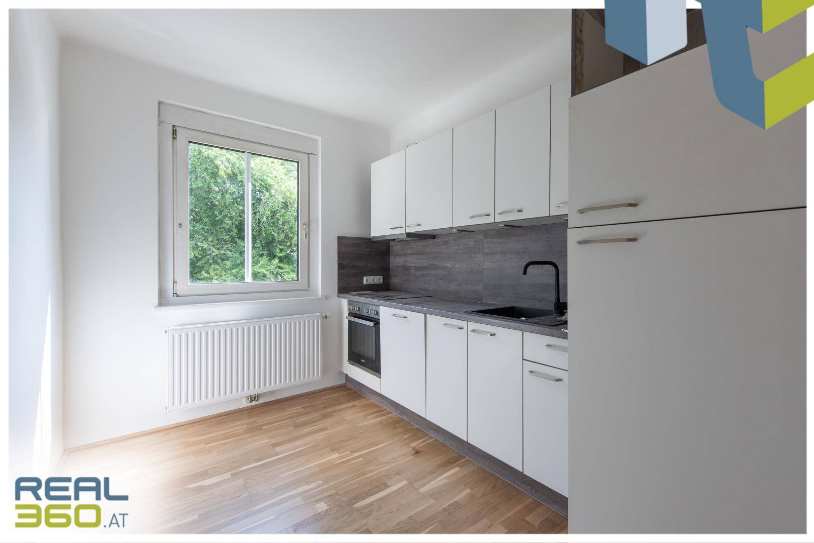 Moderne Küche mit weißen Schränken, dunkler Arbeitsplatte und Fenster mit Blick ins Grüne.