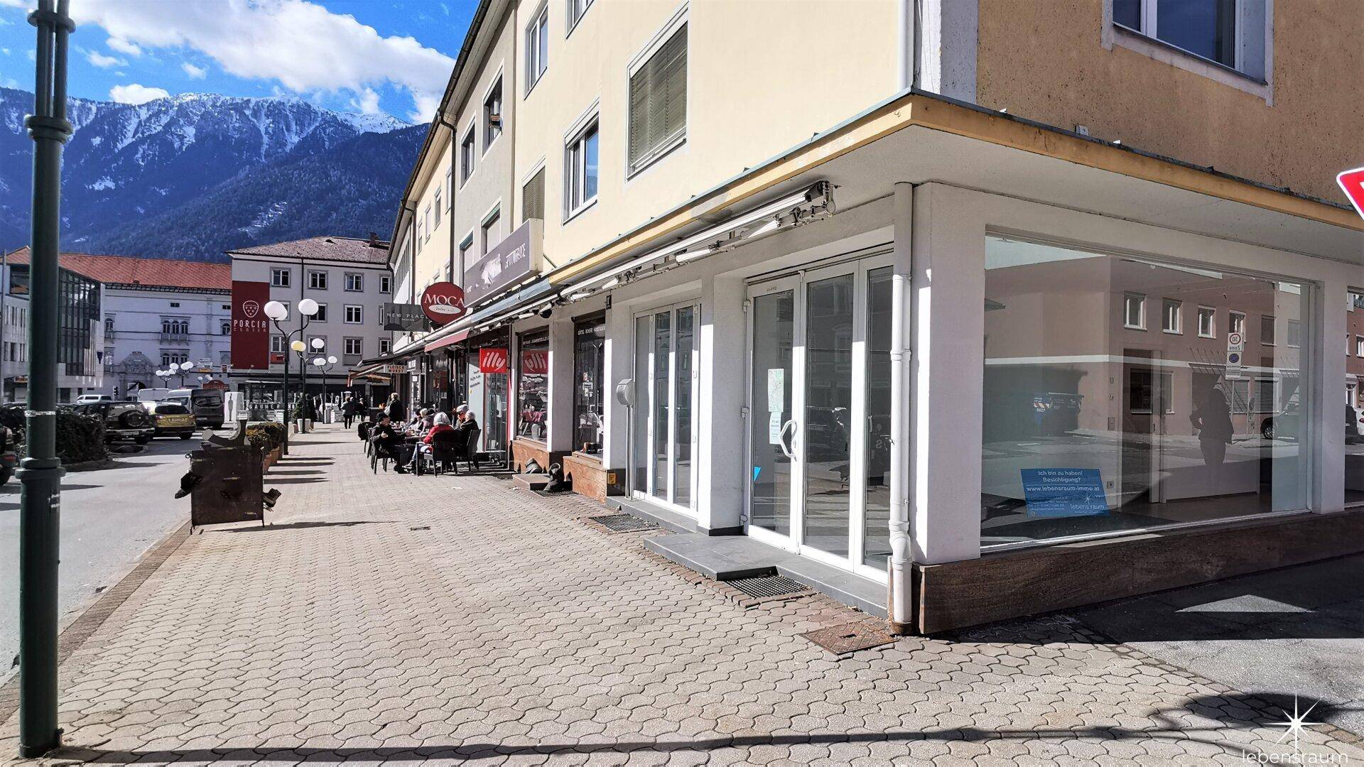 Geschäftsfassade mit großen Schaufenstern und belebtem Bürgersteig, umgeben von einer städtischen Landschaft.