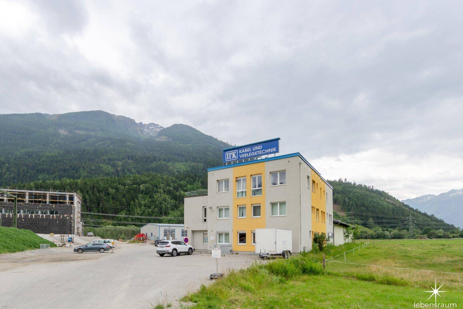 Gesamtansicht des Geschäftsgebäudes mit gelben Akzenten und umliegender Berglandschaft unter bewölktem Himmel.