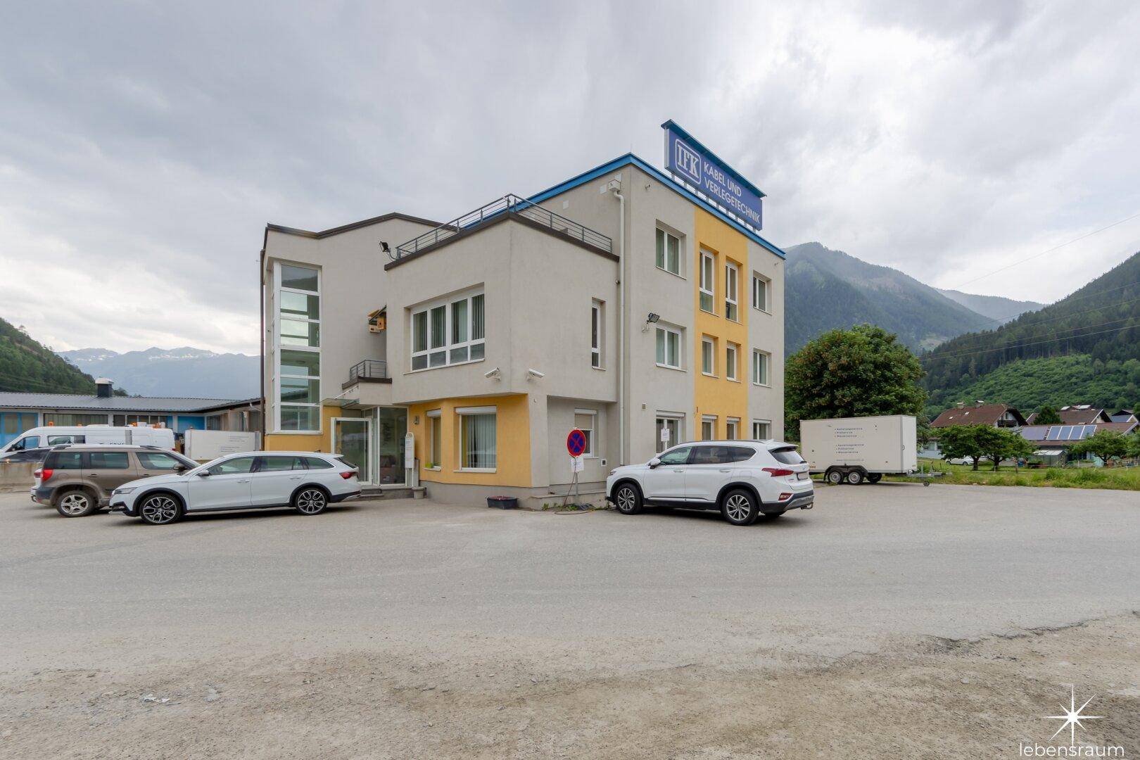 Ansicht des Bürogebäudes mit mehreren geparkten Autos und einer Bergkulisse im Hintergrund unter bewölktem Himmel.