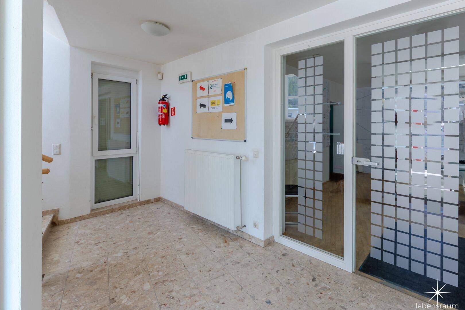 Heller Eingangsbereich mit Fliesenboden, einer Pinnwand und einer Brandschutzvorrichtung an der Wand.