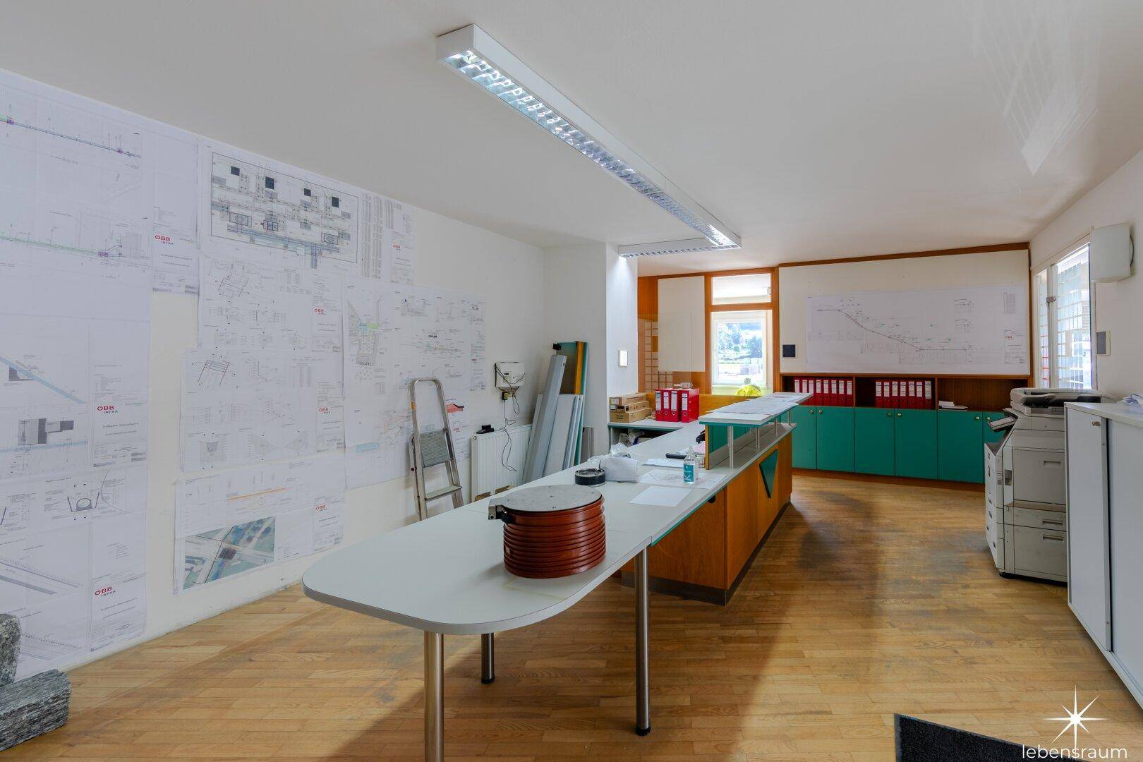 Großes Büro mit Holzboden, langen Arbeitstischen und technischen Zeichnungen an den Wänden als Dekoration.
