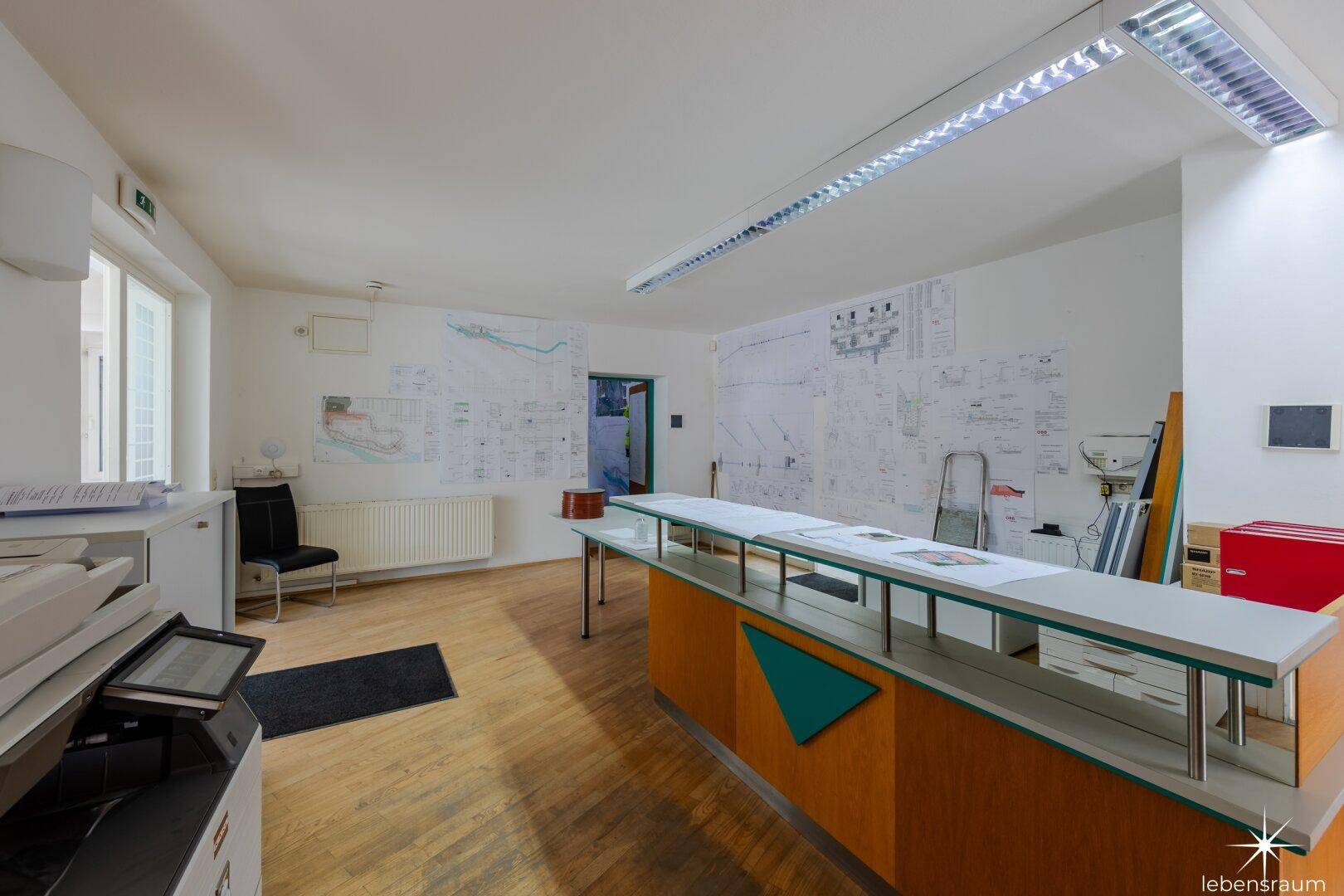 Geräumiges Büro mit Holzboden, langen Arbeitstischen und einem Drucker im Vordergrund des Raumes.