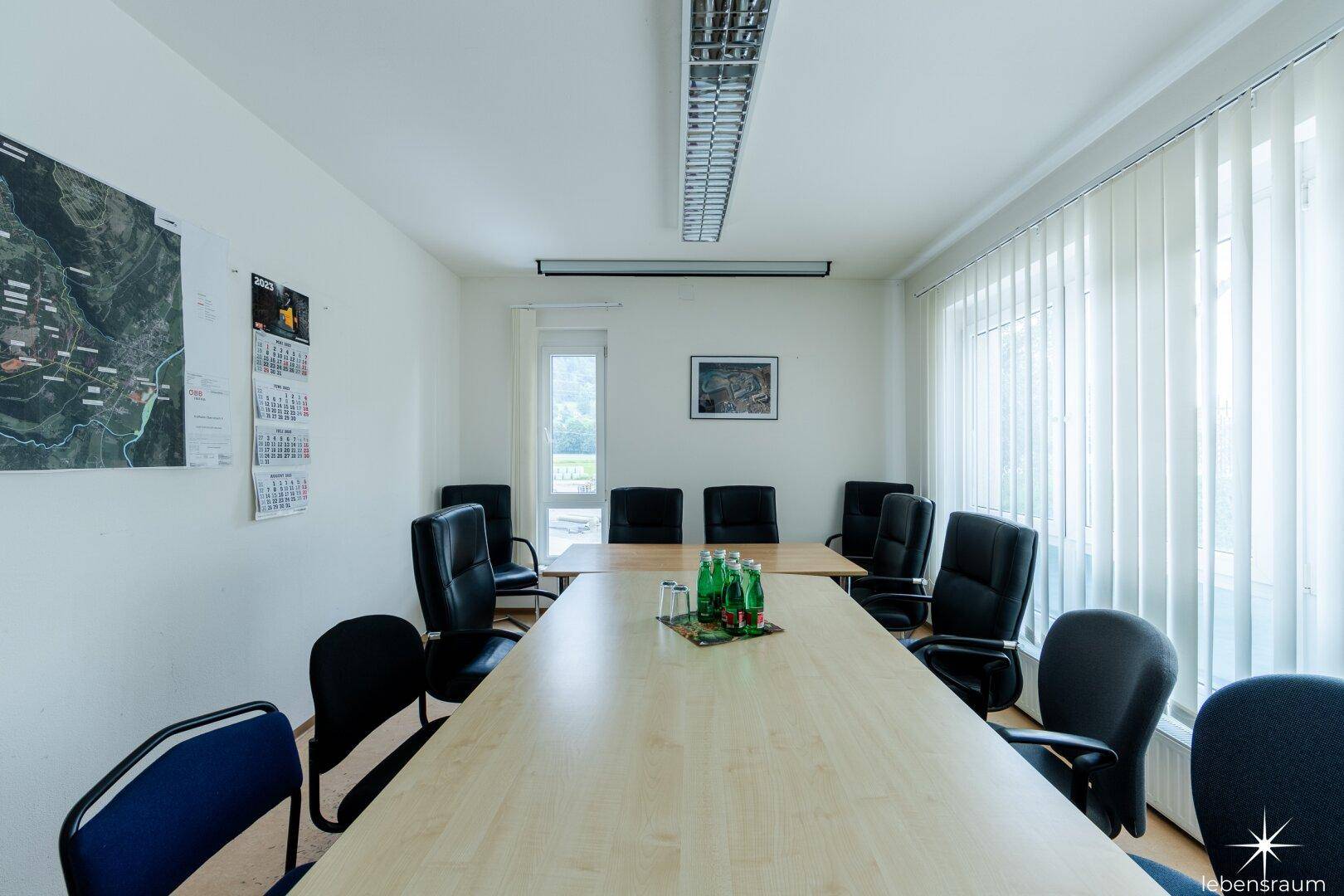 Konferenzraum mit großem Holztisch, vielen Stühlen und Fenstern mit vertikalen Jalousien.