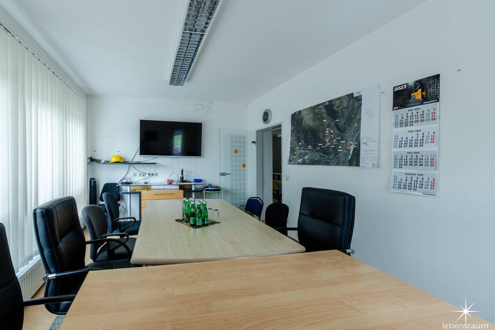 Besprechungsraum mit großem Tisch, Stühlen und einem an der Wand montierten Fernseher.