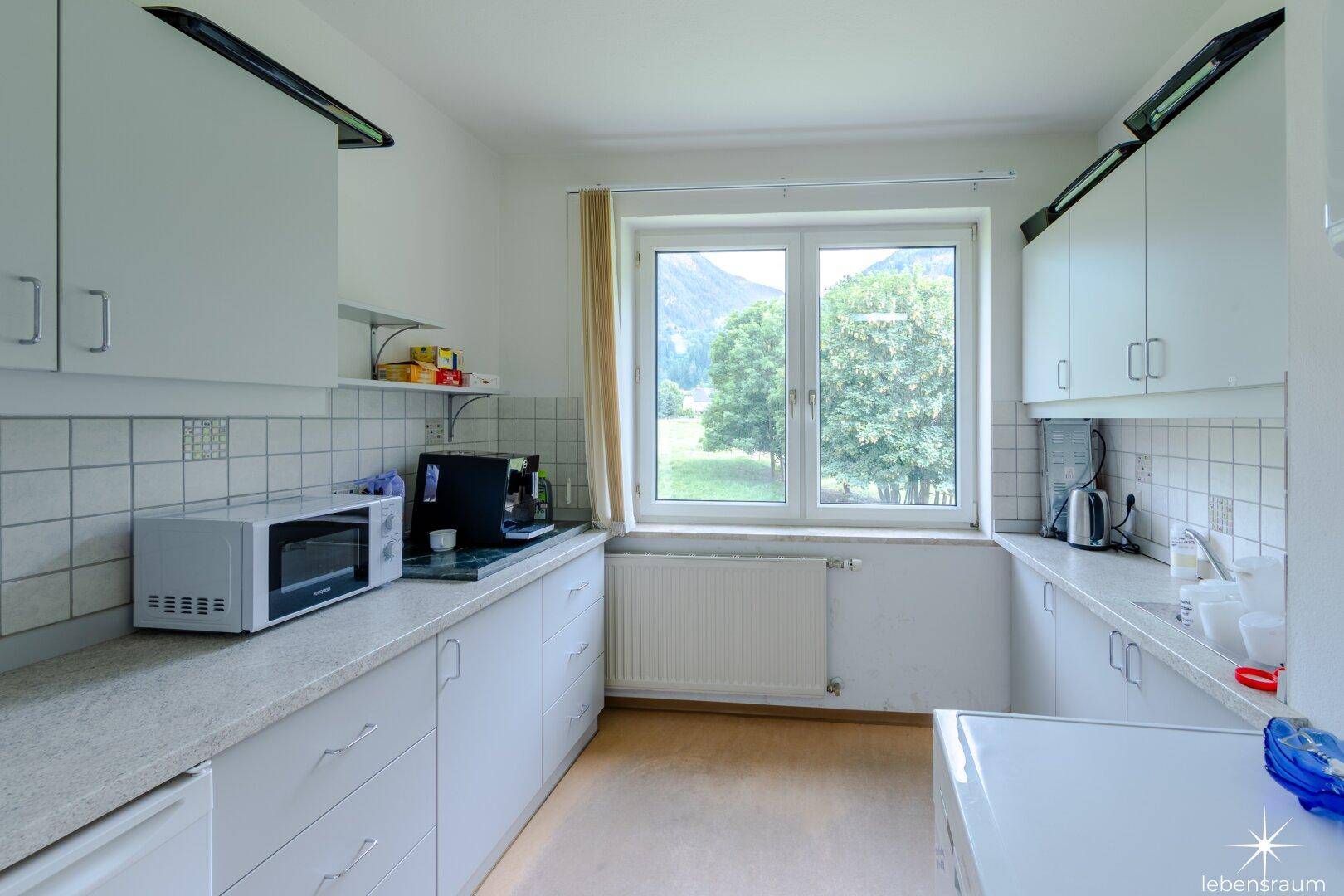 Funktionale Küche mit weißen Einbauschränken, Mikrowelle und einem großen Fenster mit Aussicht.