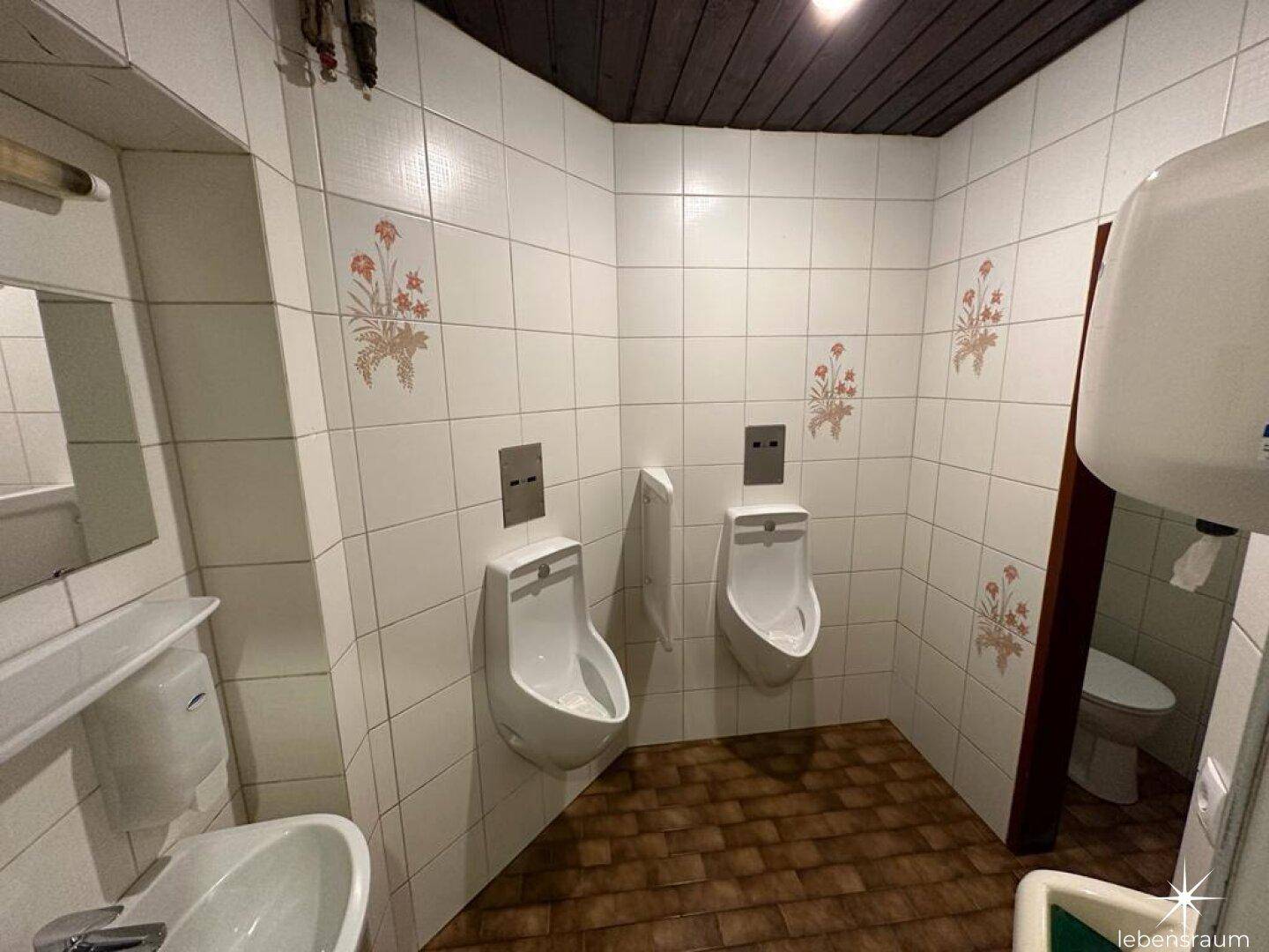 Herren-Gästetoilette mit zwei Urinalen, Waschbecken und weißen Fliesen.