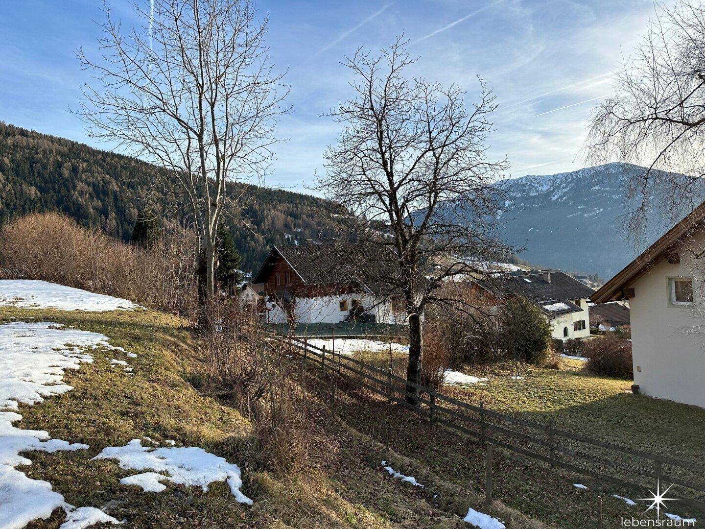 Hanglage mit kahlen Bäumen, Schneeresten und Häusern im Hintergrund, umgeben von Bergen.