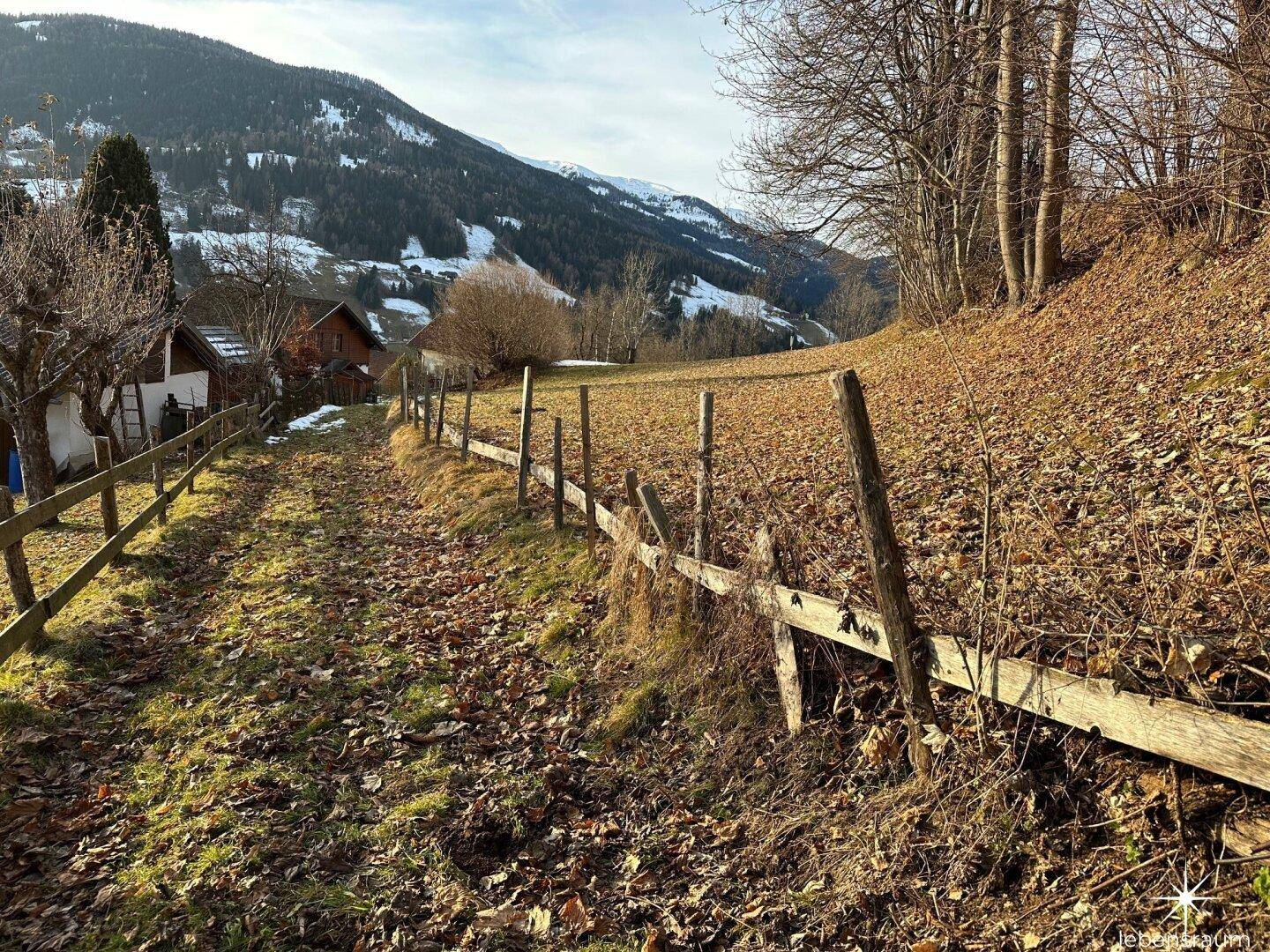 Ländlicher Weg mit rustikalem Holzzaun und Blick auf die bewaldete Berglandschaft.