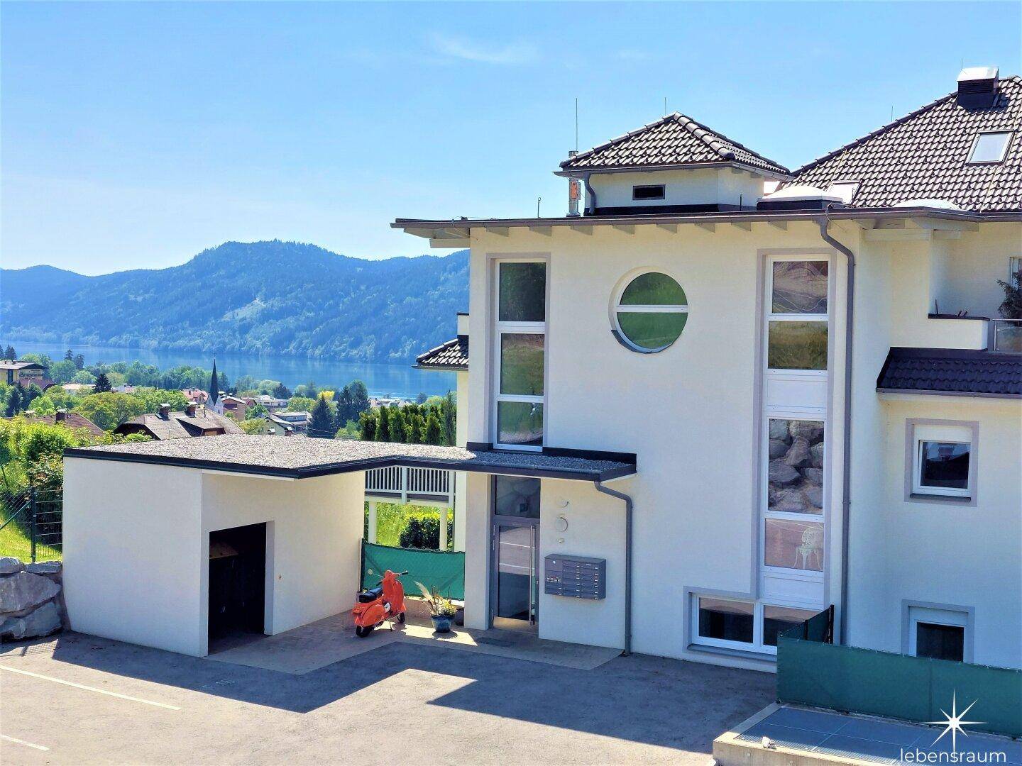 Moderne Gebäudefassade mit großen Fenstern und Carport, mit Seeblick im Hintergrund.