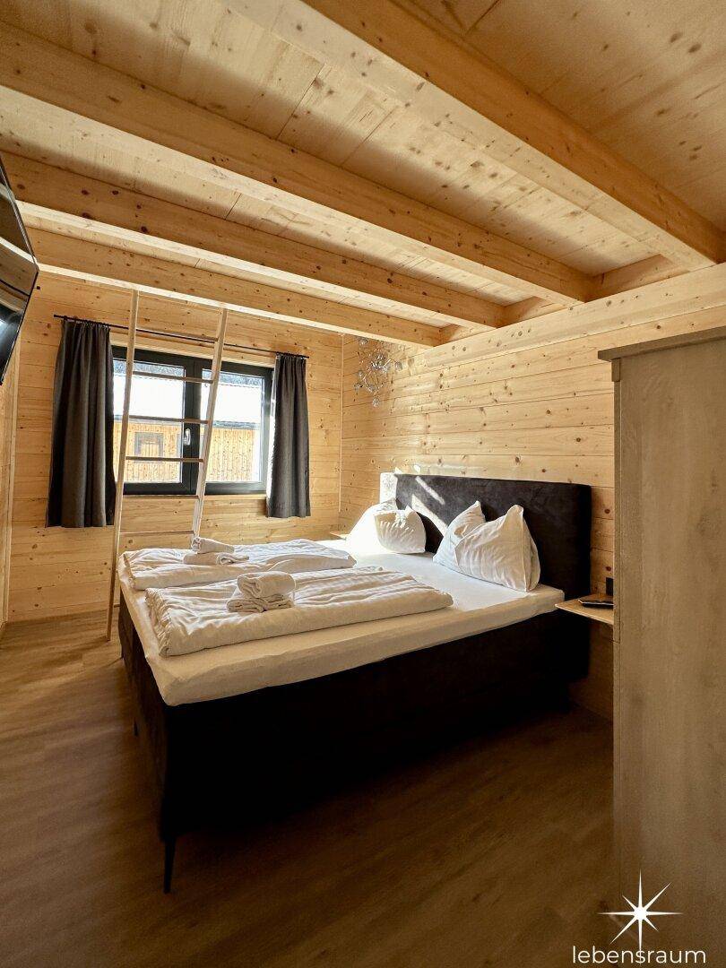 Schlafzimmer mit Doppelbett, Holzwänden, Dachschräge und Fenster mit Blick nach draußen.