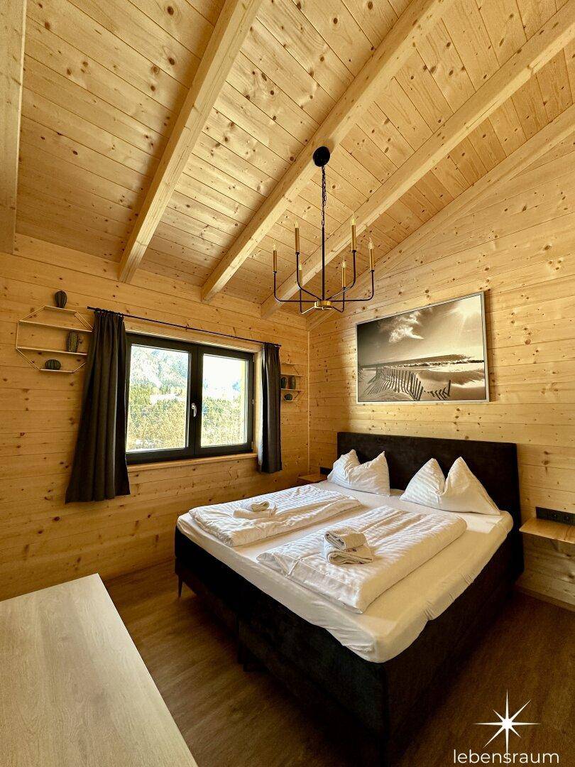 Helles Schlafzimmer mit Doppelbett, Holzwänden, Dachschräge und modernem Kronleuchter.