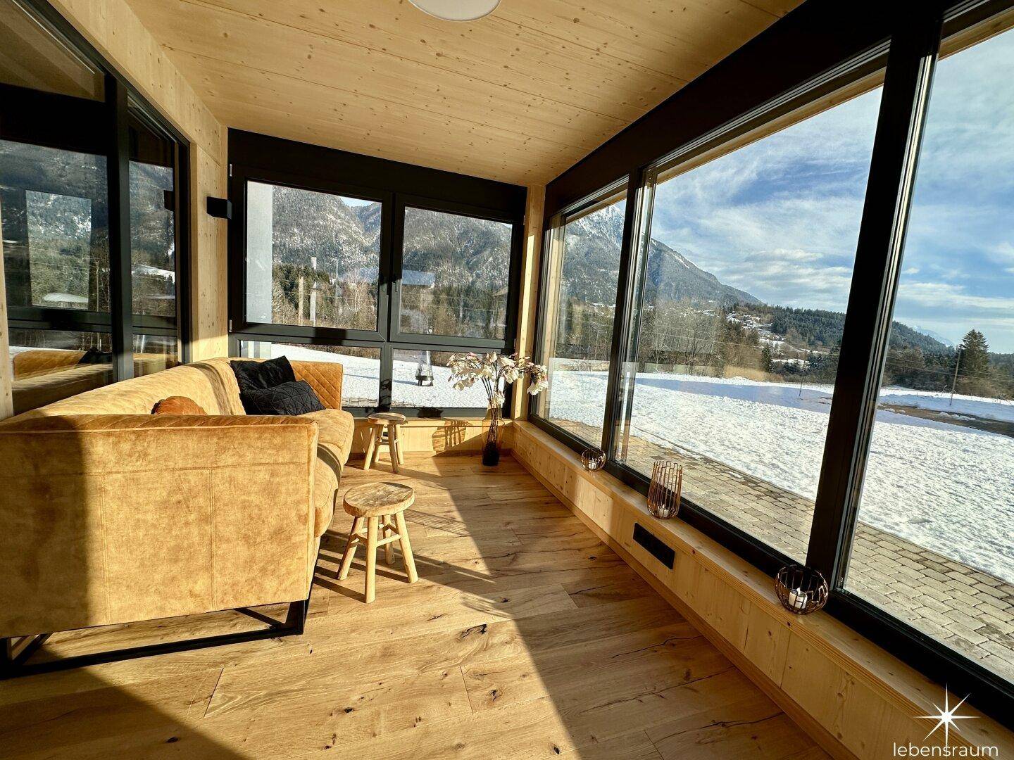 Lichtdurchfluteter Wintergarten mit Sofa und Panoramafenstern mit Blick auf die Winterlandschaft.