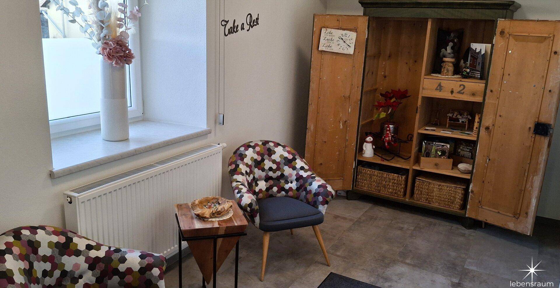 Gemütlicher Loungebereich mit einem farbenfrohen Sessel, einem Holzschrank und einem Fenster mit Heizkörper.