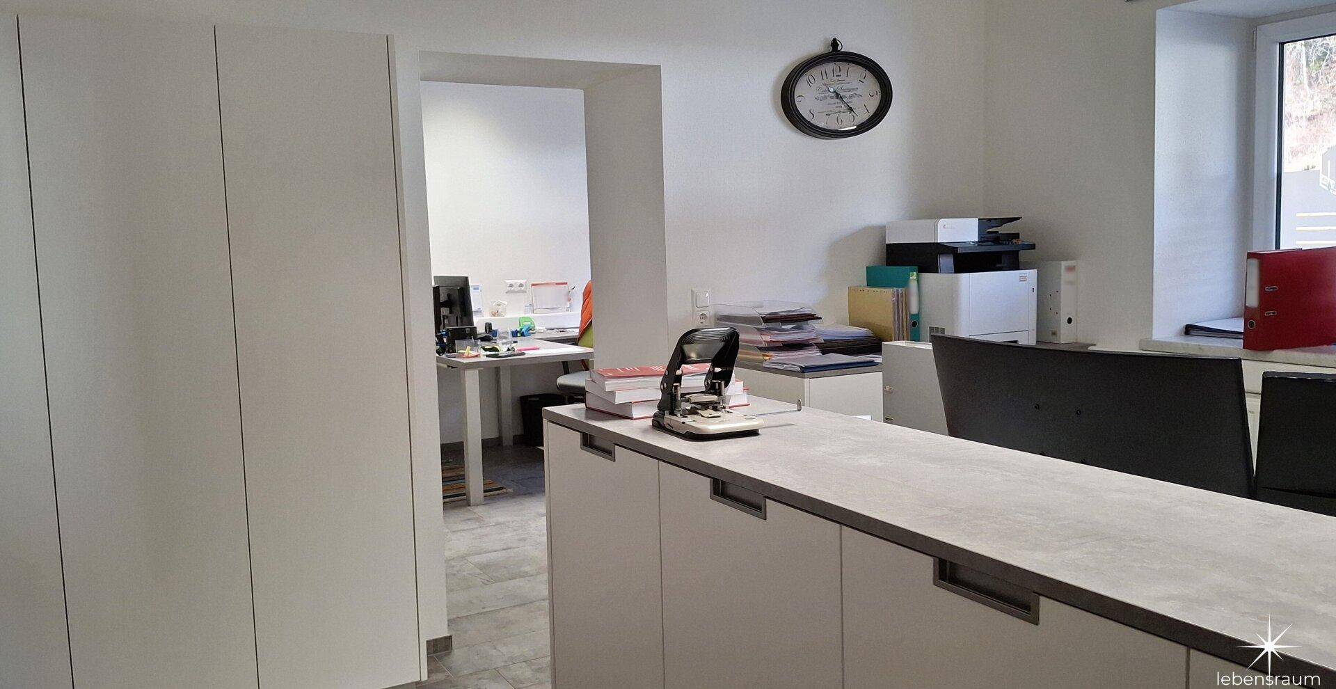 Großzügiger Büroraum mit weißen Schränken und einem Durchgang zu einem weiteren Arbeitsbereich.