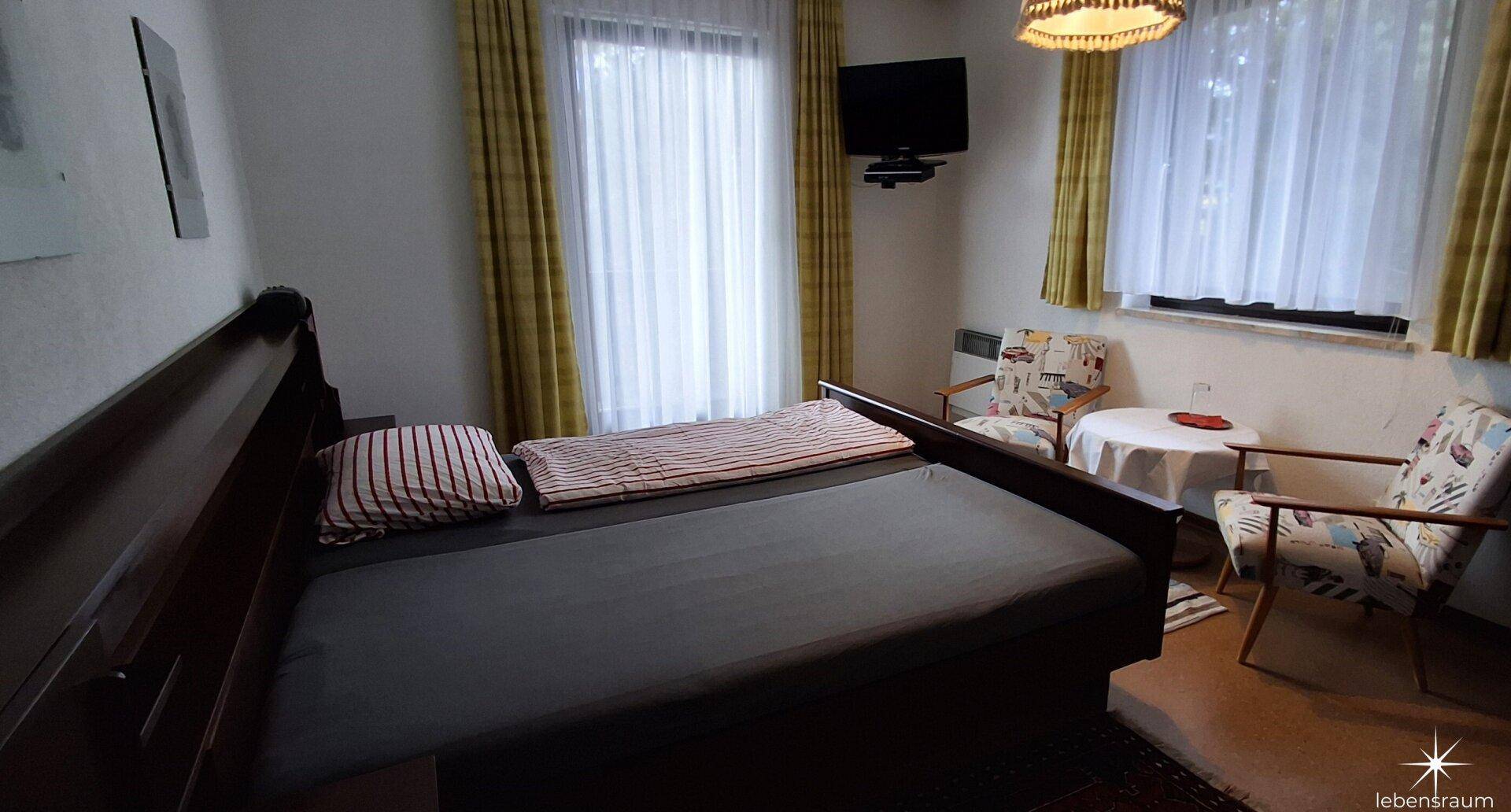Schlafzimmer mit Doppelbett, Fernseher und Vorhängen an den Fenstern.