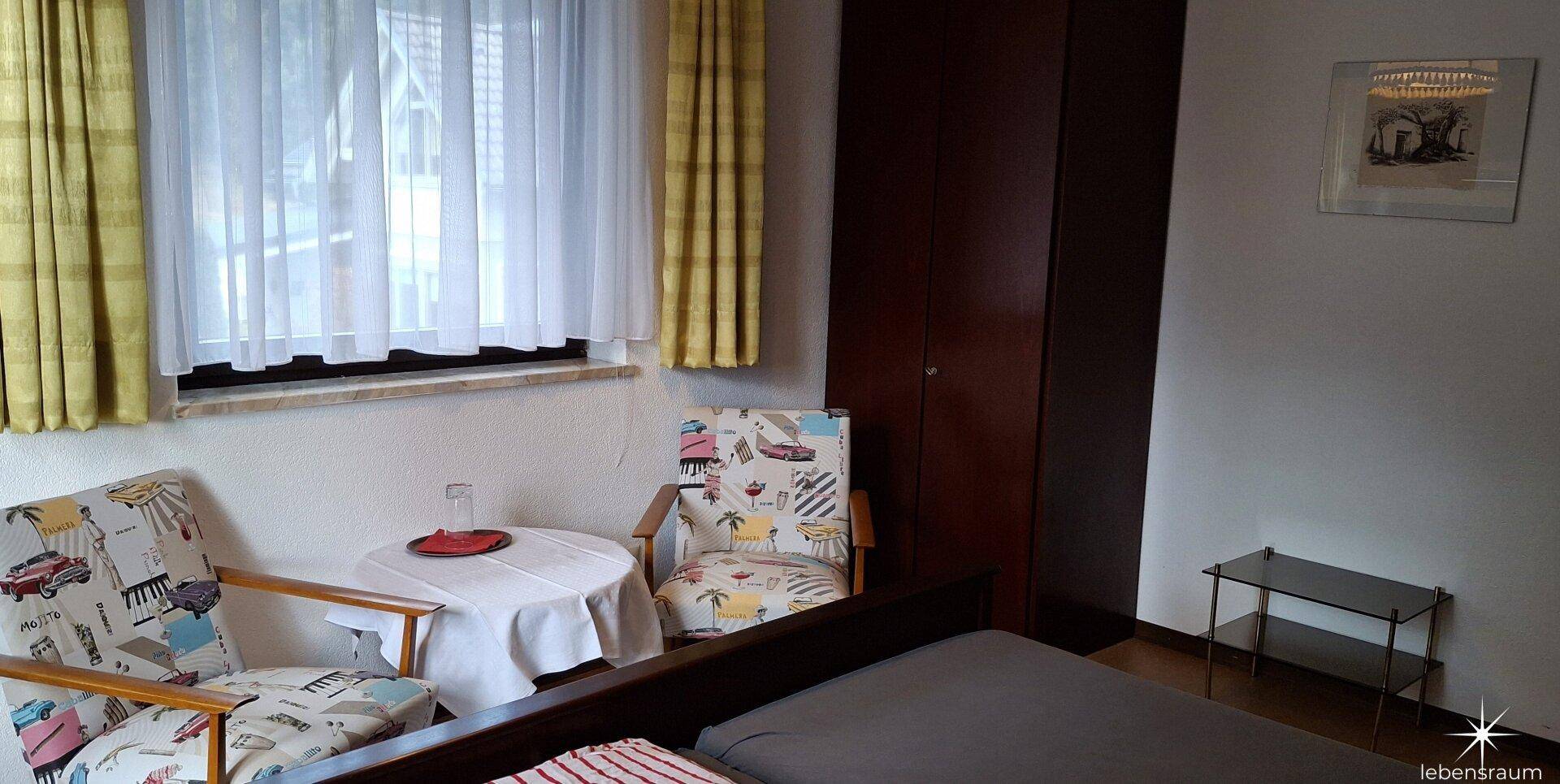 Helles Schlafzimmer mit Kleiderschrank, zwei Sesseln und Fenster.