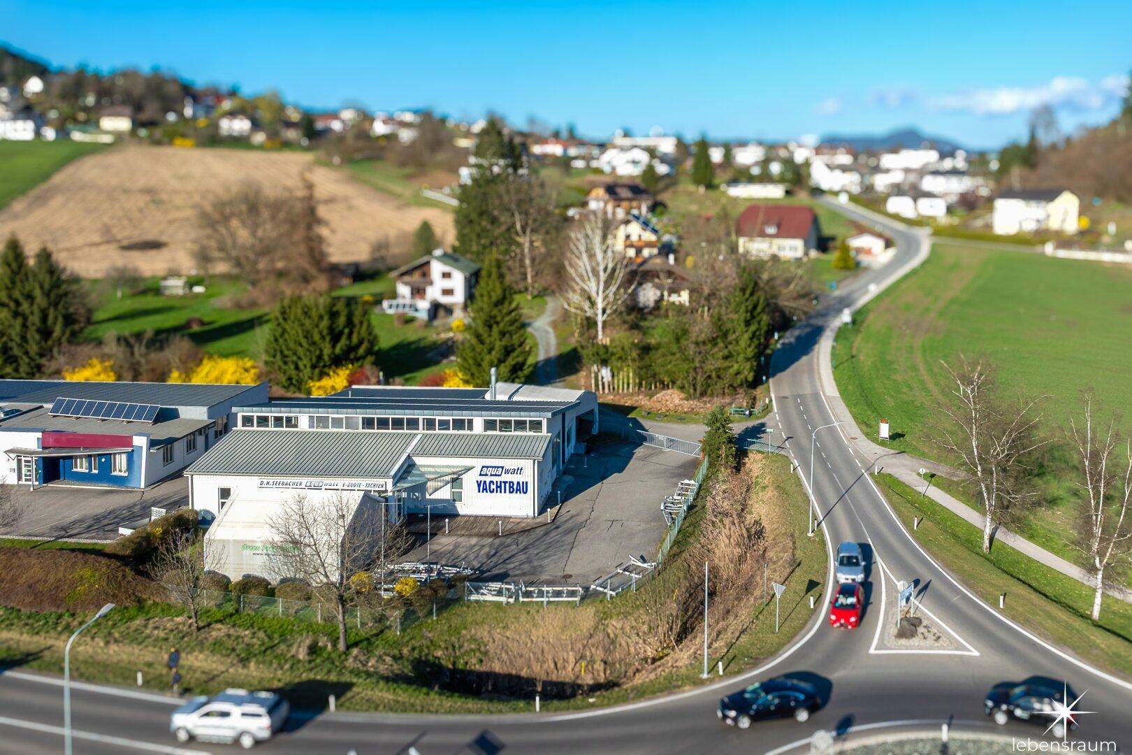 Weitläufige Luftaufnahme des Gebäudes mit angrenzender Straße, Kreisverkehr und ländlicher Umgebung unter klarem Himmel.