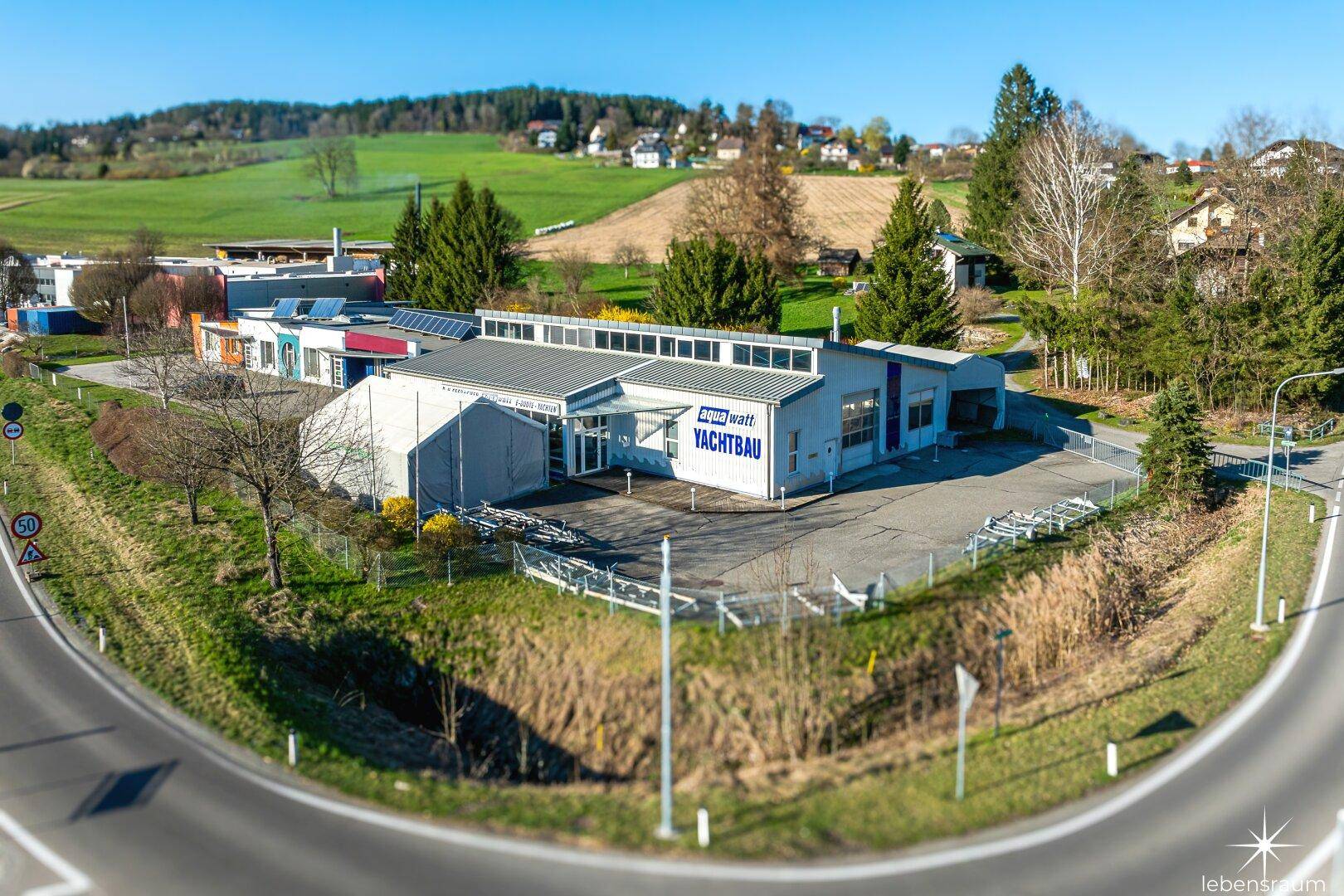 Luftaufnahme des Gebäudes mit umliegenden Grünflächen, einer Straße und einem Kreisverkehr im Vordergrund bei Sonnenschein.