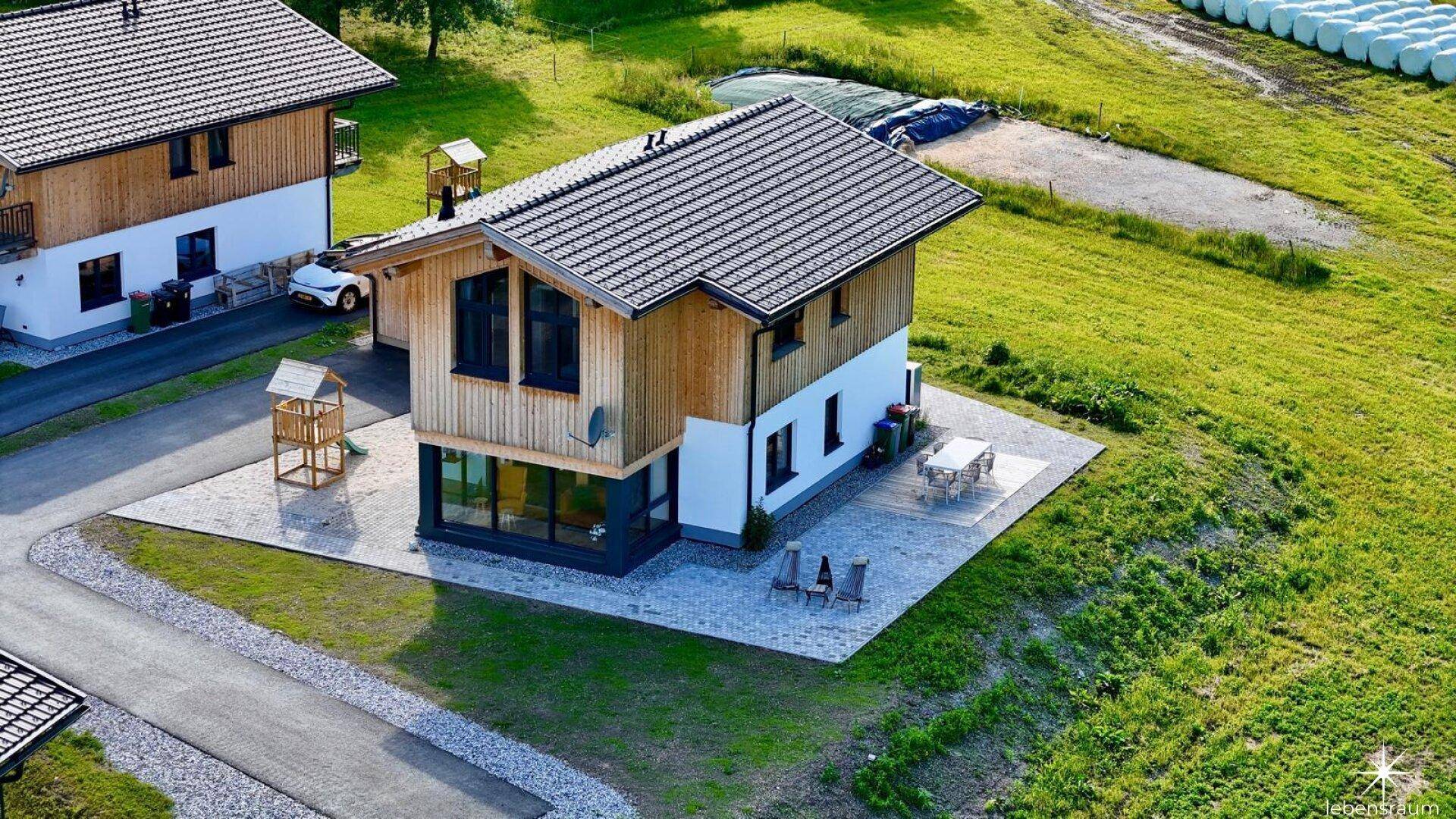 Modernes Haus mit Holzfassade und Terrasse in ländlicher Umgebung, umgeben von grünen Wiesen.