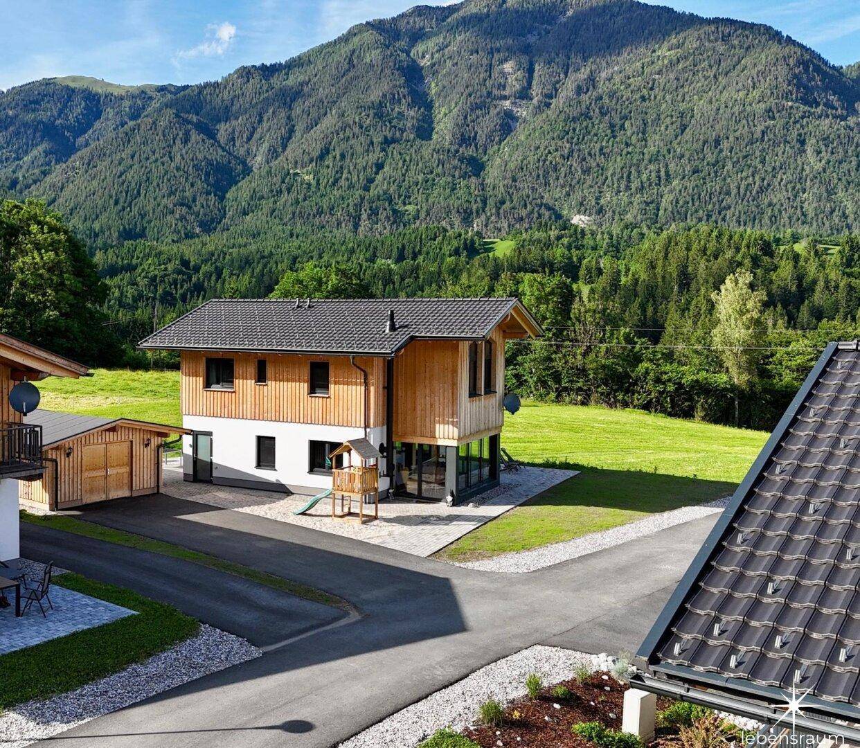 Modernes Wohnhaus mit Holzfassade und Spielplatz, umgeben von grünen Wiesen und majestätischen Bergen.