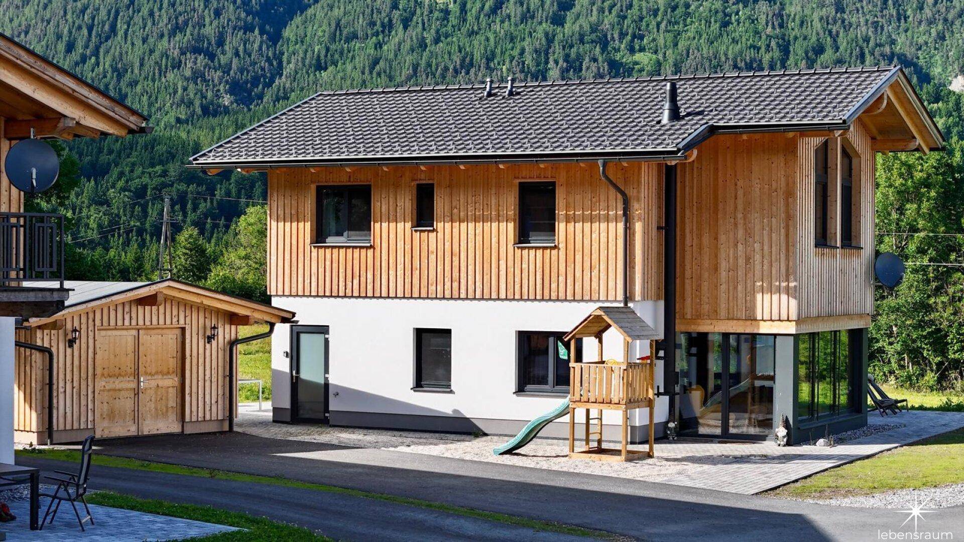 Detailansicht des modernen Hauses mit heller Holzfassade, großen Fenstern und einem kleinen Spielplatz.