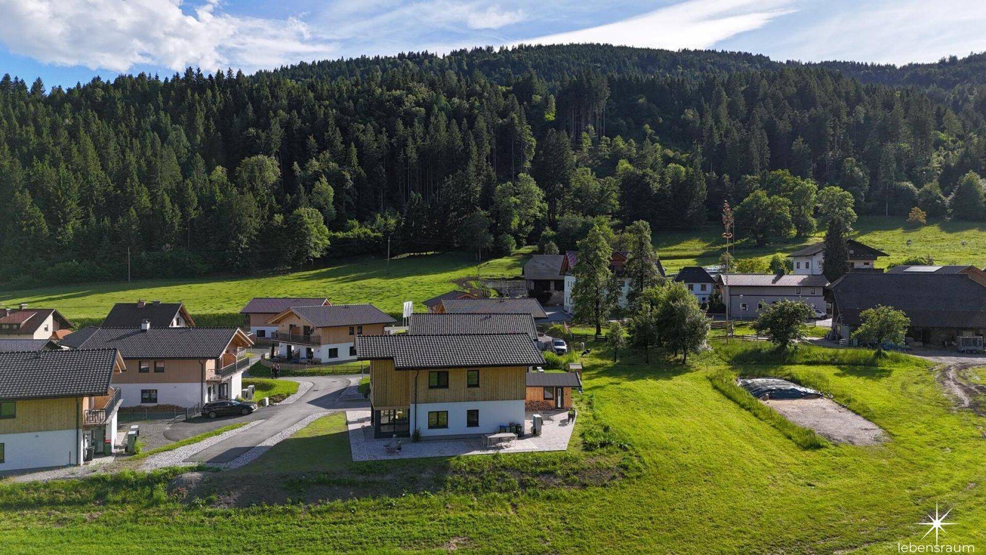 Siedlung moderner Häuser in ländlicher Umgebung, eingebettet in grüne Wiesen und bewaldete Hügel.