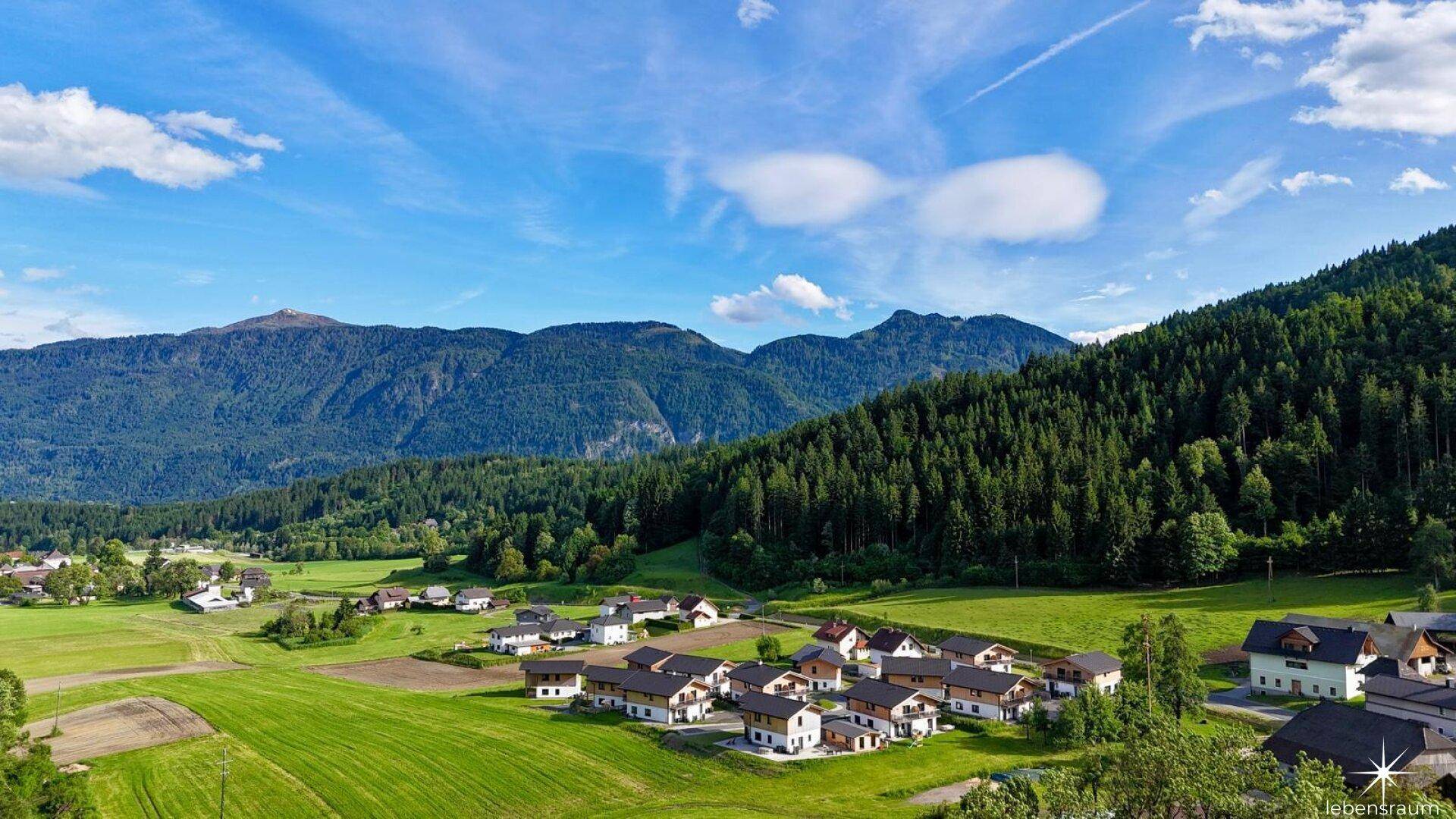 Weitläufige Landschaft mit einer Siedlung, umgeben von grünen Feldern und einer beeindruckenden Bergkette.