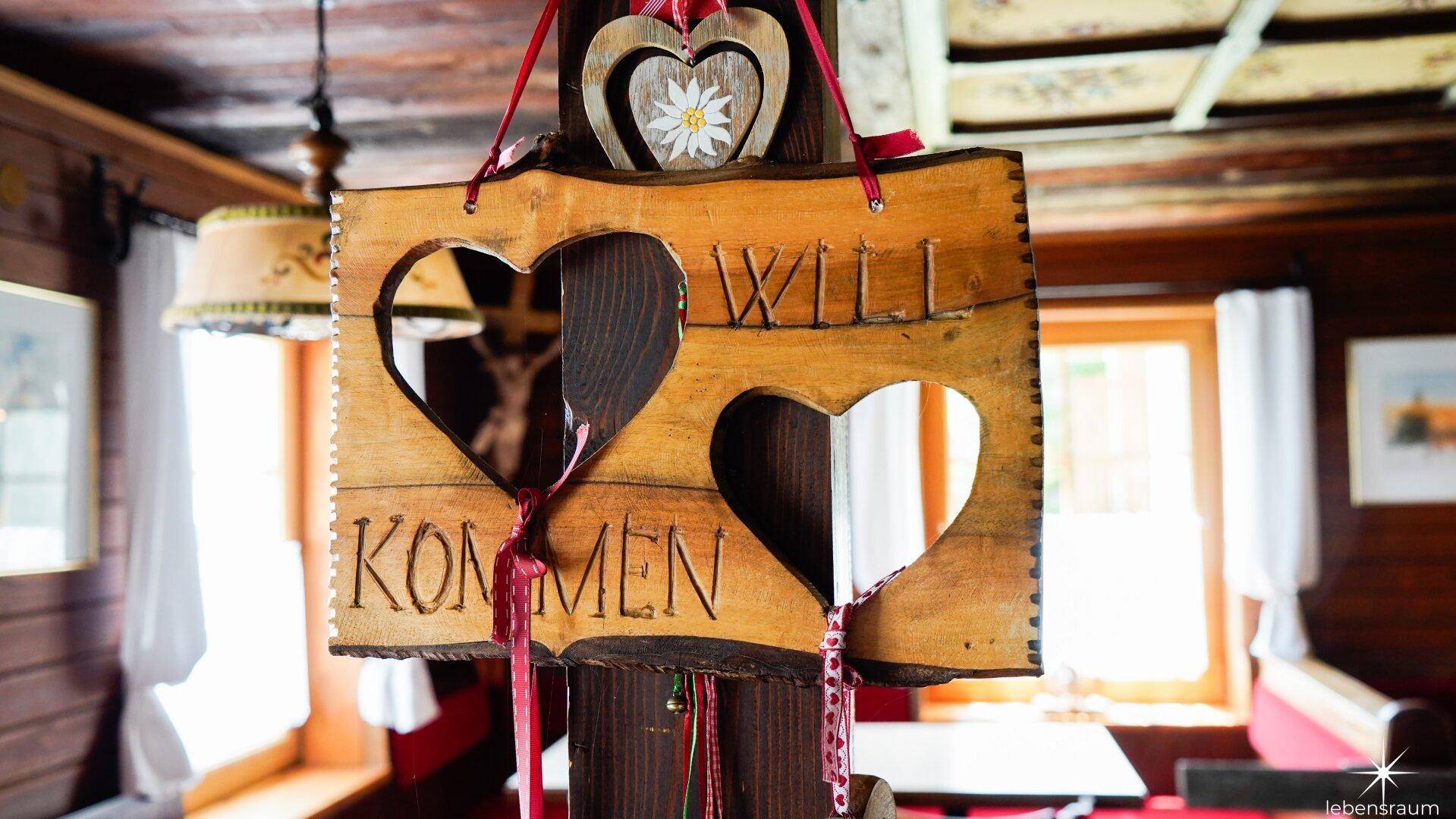 Rustikales Holzschild mit Herzmotiven und der Aufschrift 'Willkommen' in einem traditionellen Raum.