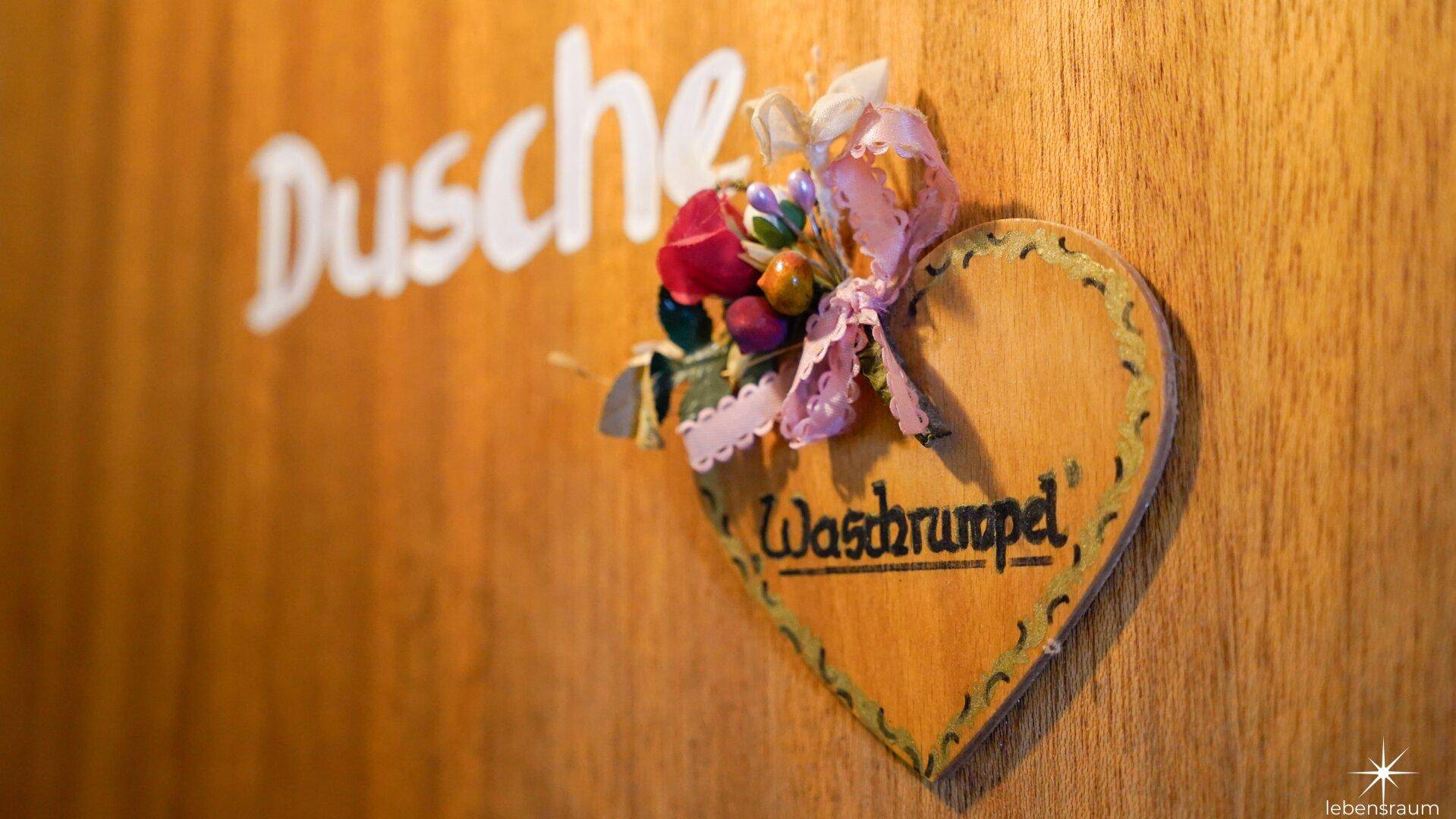 Dekoratives Holzschild mit Aufschrift 'Dusche' und Blumenverzierung an einer Holzwand.